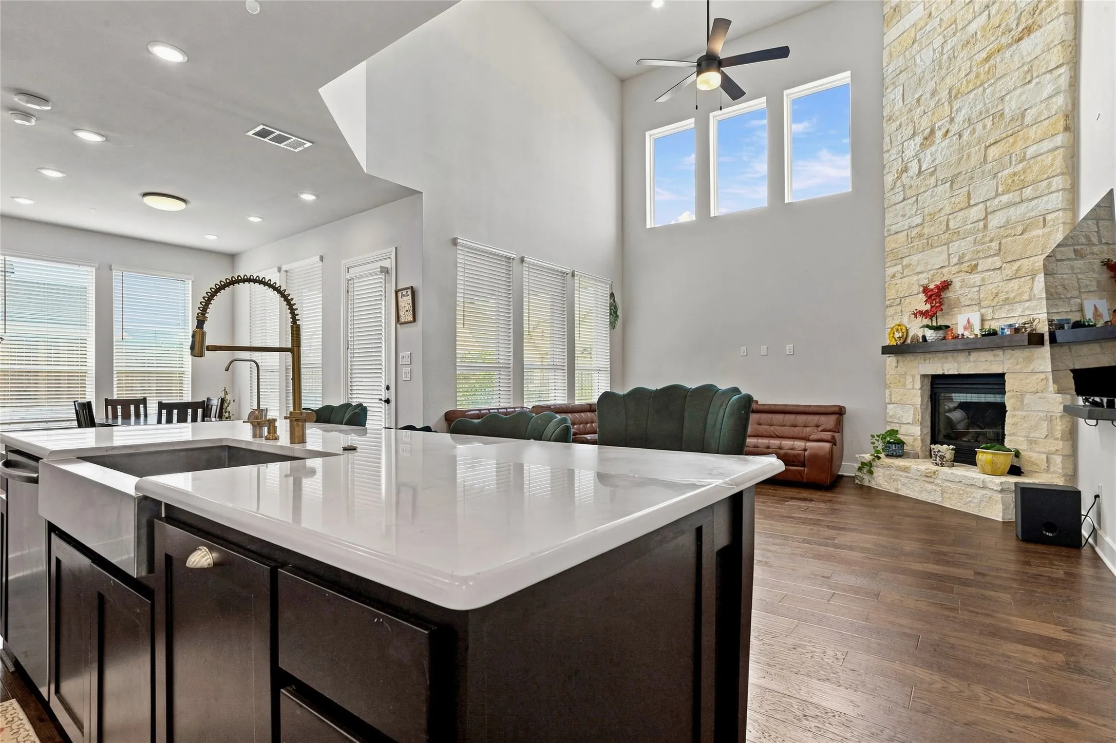 Spacious kitchen island with white countertop and farmhouse sink, dark wood-finish cabinetry, wood-finish flooring, stone fireplace with mantel, and high ceilings with multiple windows