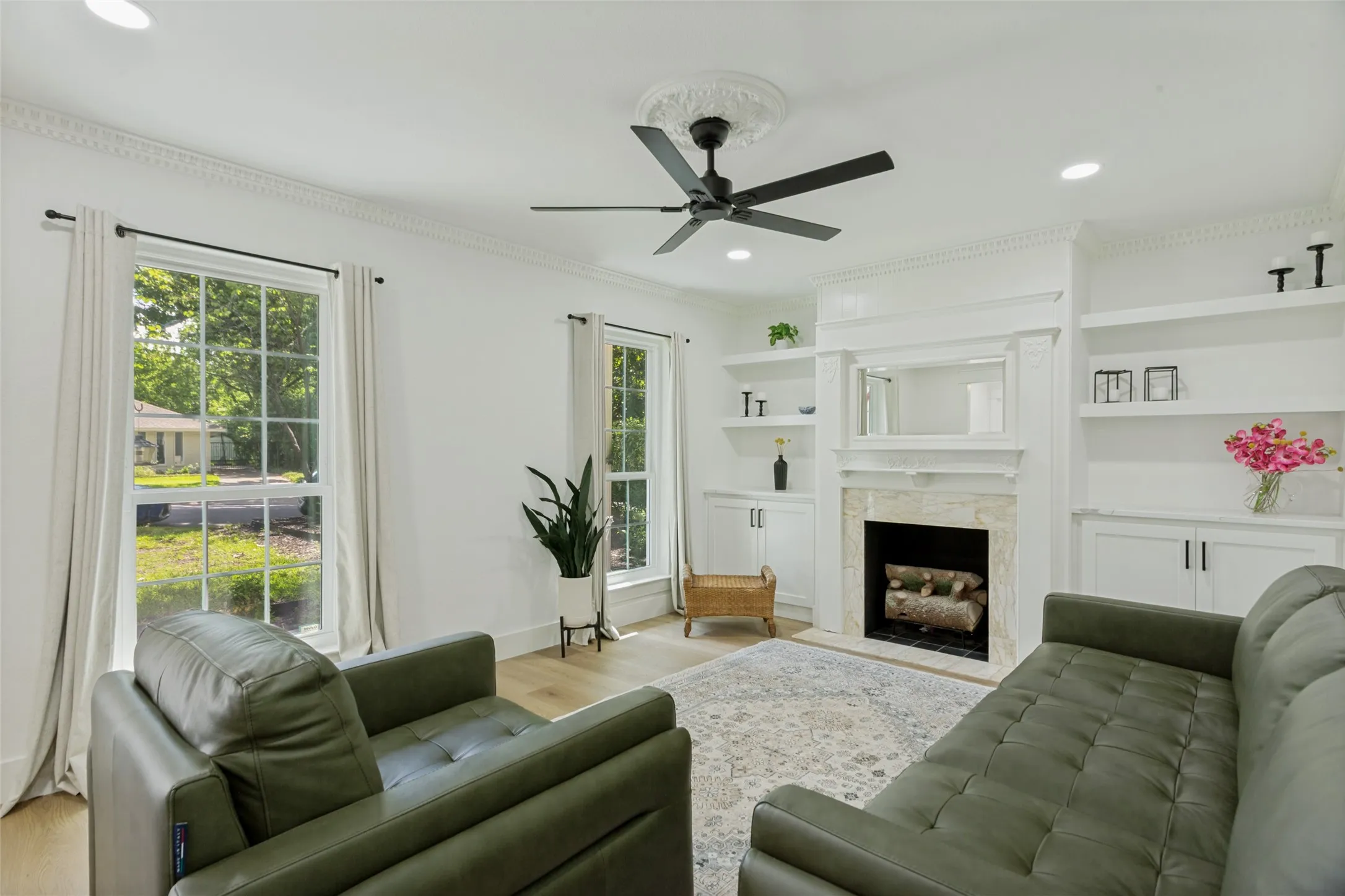 Living area featuring light wood-finish flooring, a ceiling fan with decorative medallion, a fireplace with stone surround, and built-in shelving and cabinetry