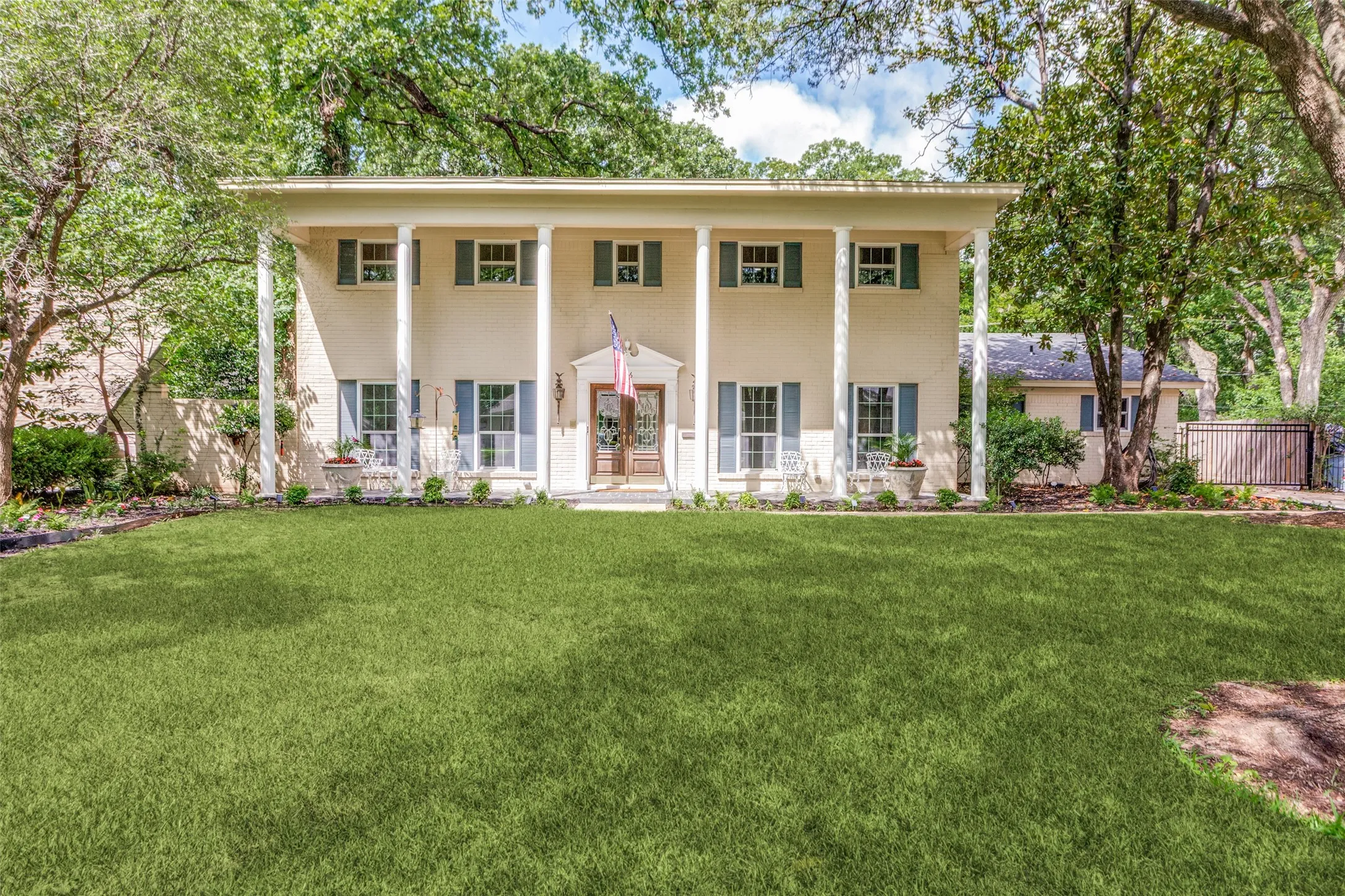 Two-story Colonial-style exterior, full-width covered porch with white columns, light-colored brick facade, dark-toned window shutters, and a prominent front lawn