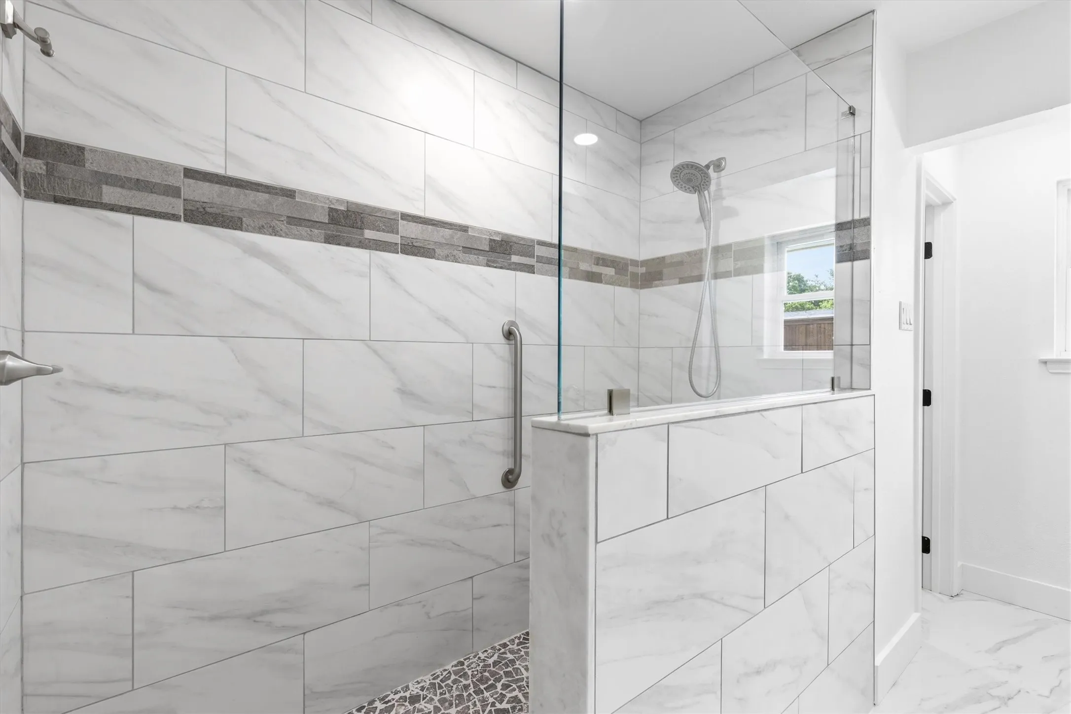 Shower enclosure featuring large format white tile with grey veining, a decorative grey tile accent band, and a glass partition