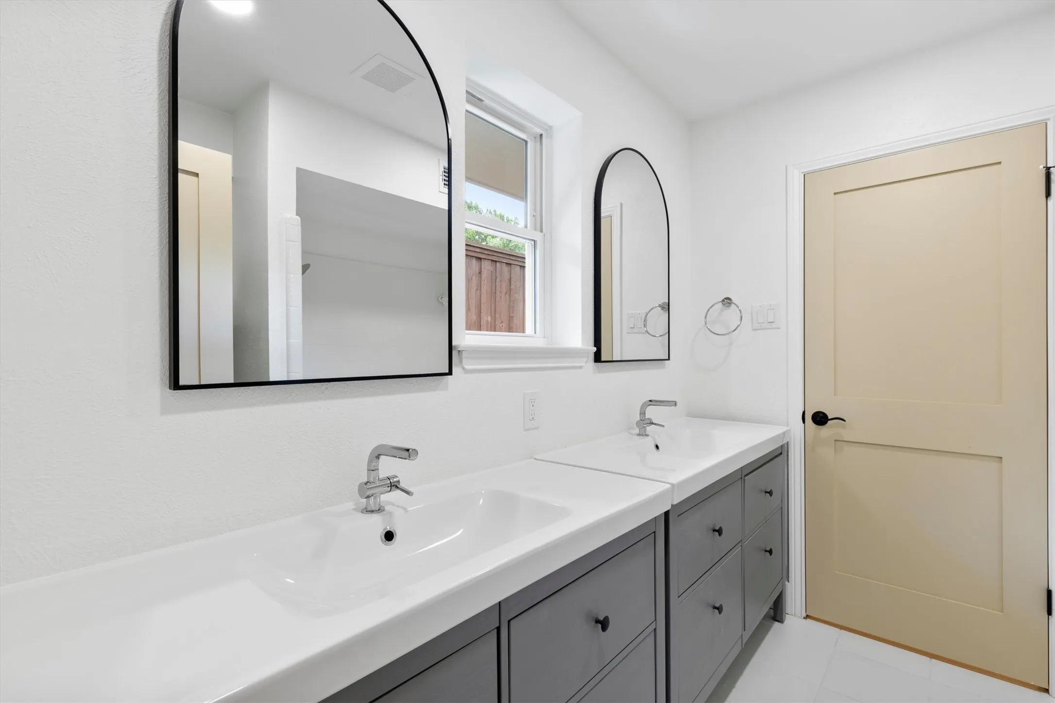 Dual vanity with integrated sinks, modern arched mirrors, chrome faucets, gray cabinetry, and a window