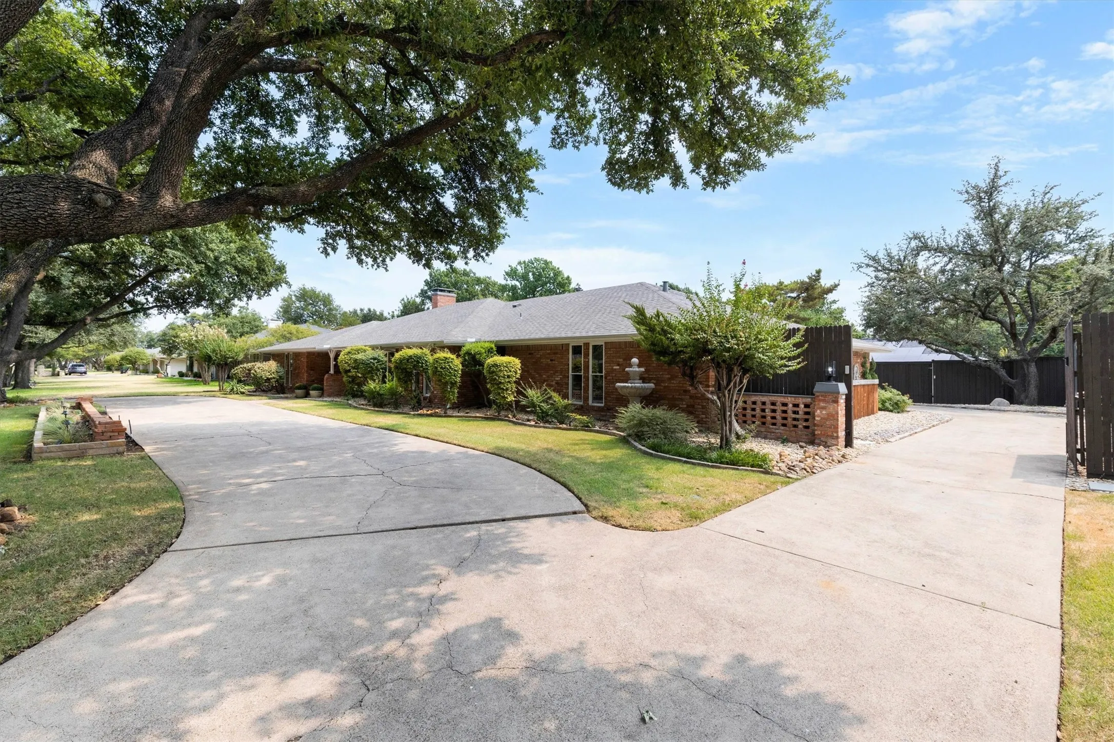 Brick exterior, mature trees, and a concrete driveway with fence and electronic gate
