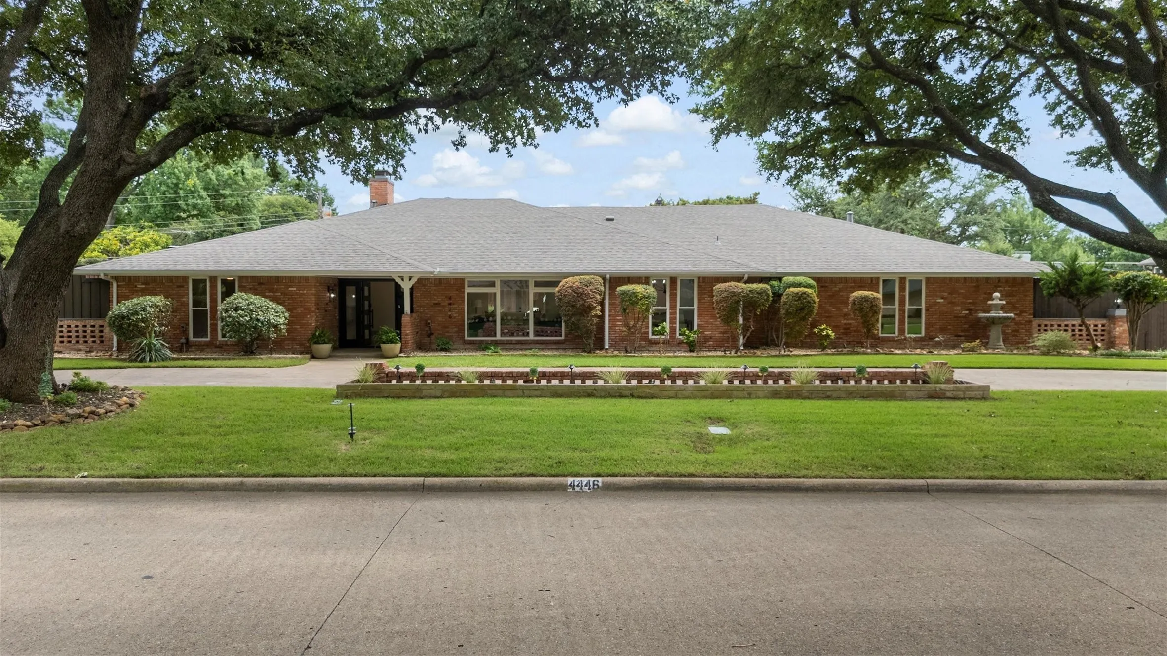 Brick facade exterior with a gabled roof, mature trees, and a manicured lawn
