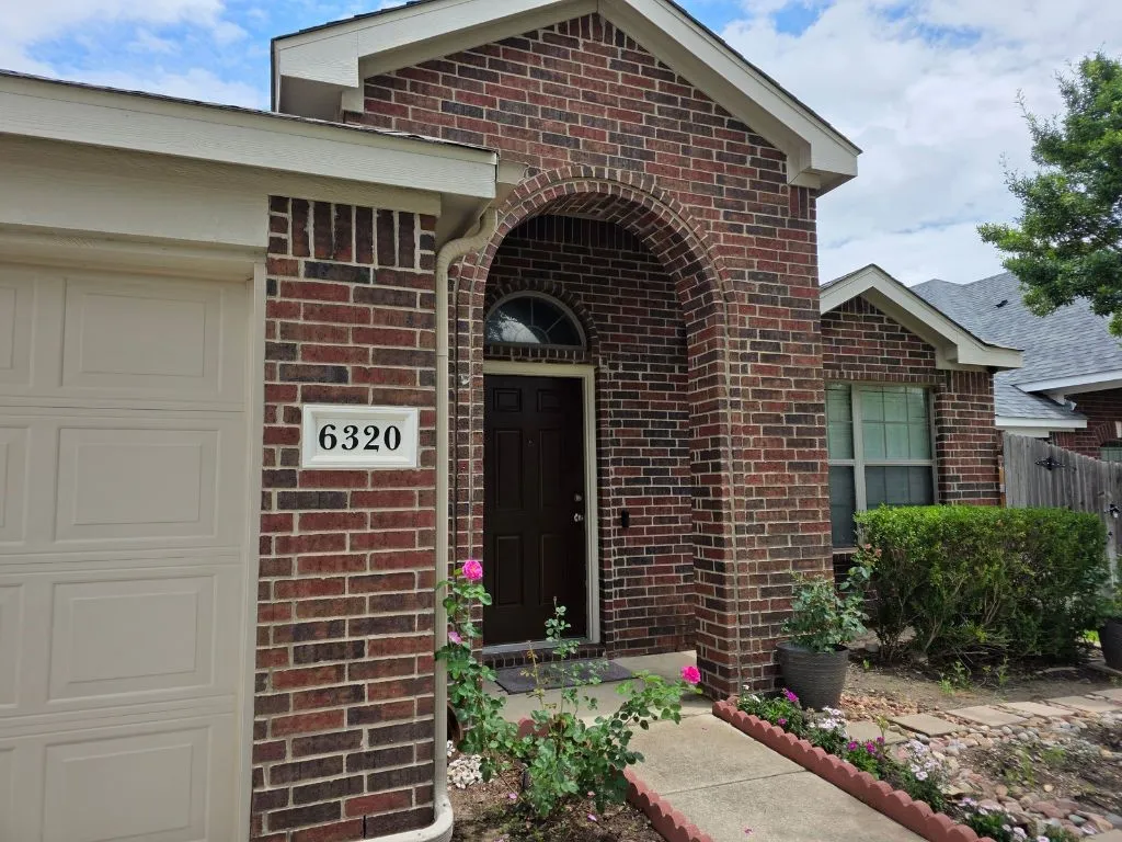 Brick exterior featuring an arched entryway, a paneled garage door, a covered porch, a concrete walkway, and established landscaping