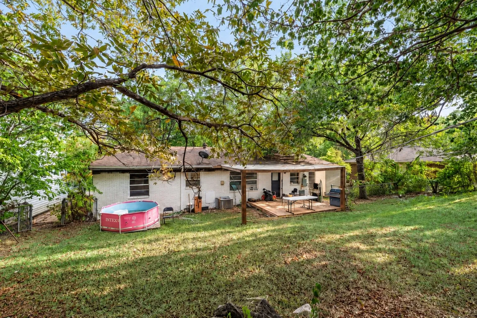 Expansive backyard featuring a partially covered deck, white brick exterior, and abundant mature trees