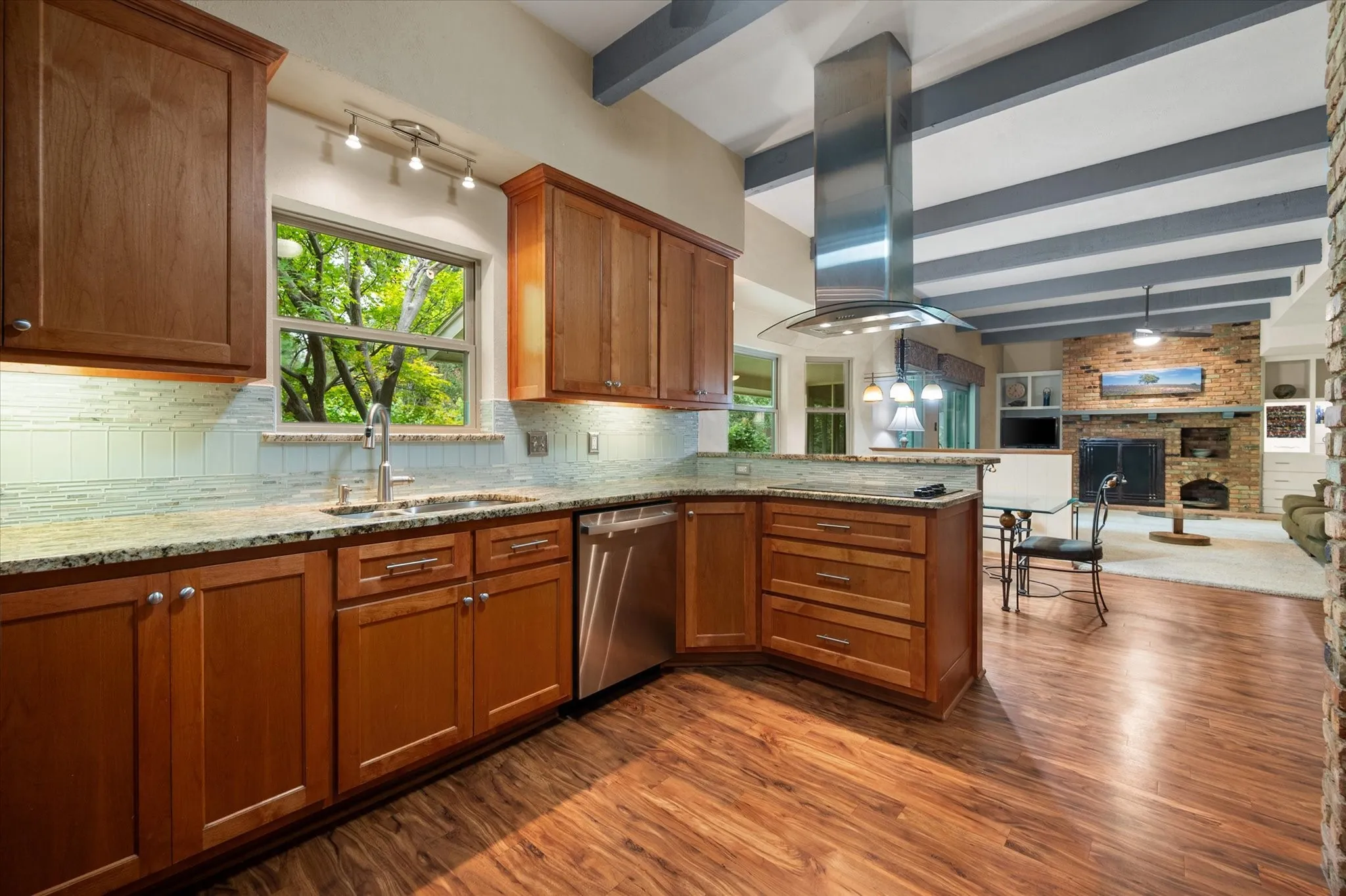 Kitchen featuring wood-finish flooring, wood cabinetry with under-cabinet lighting, a stainless steel range hood, a built-in dishwasher and views over the kitchen sink of the wooded backyard.