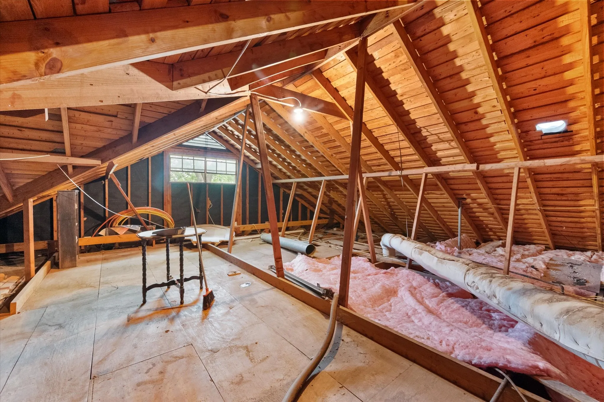 Spacious unfinished attic space featuring exposed wooden rafters, a gabled window, and plywood flooring