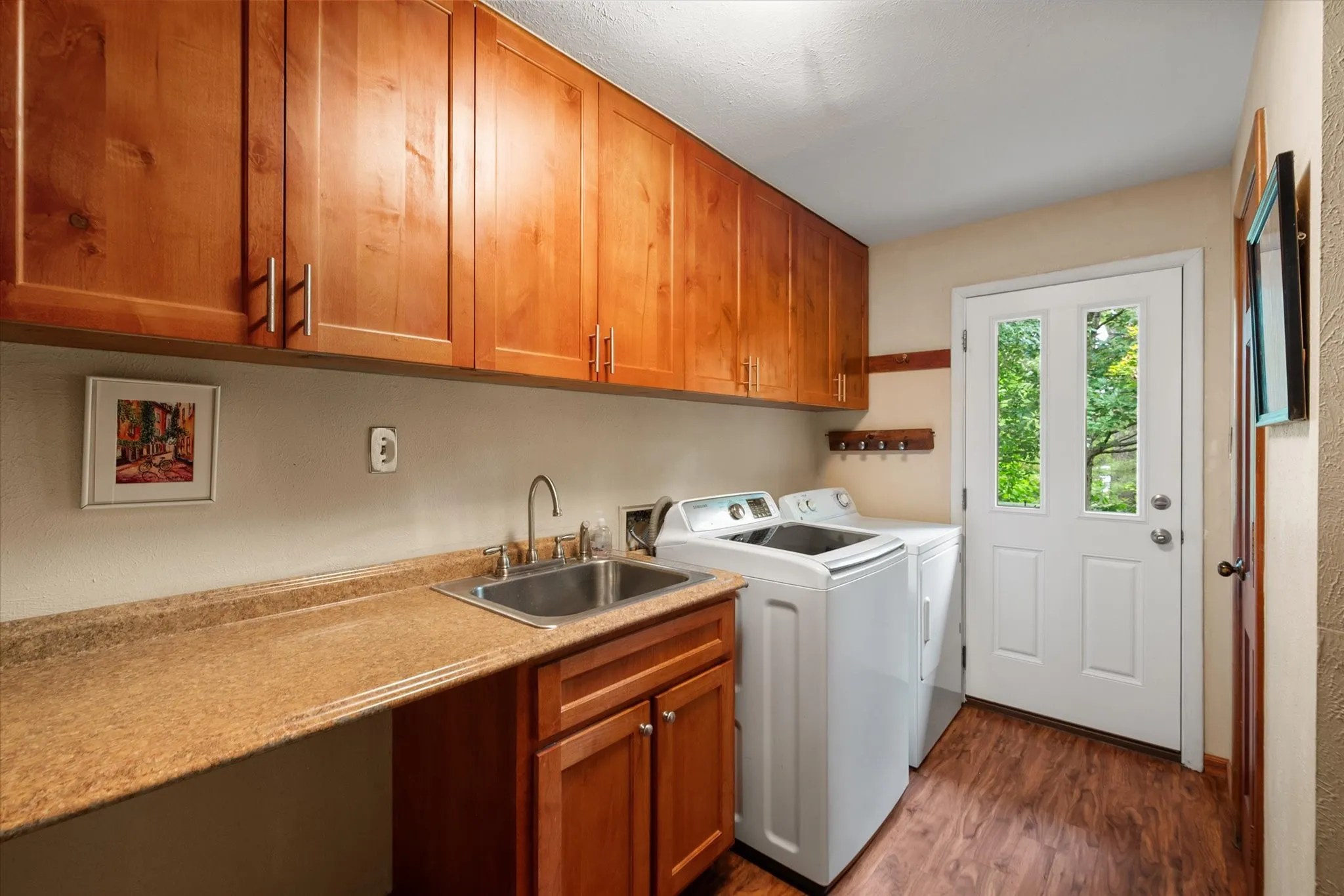 Laundry room features expansive upper and lower wood cabinetry, stainless steel sink, lots of counter space, wood-finish flooring, and a glass-paneled exterior door leading to the backyard.