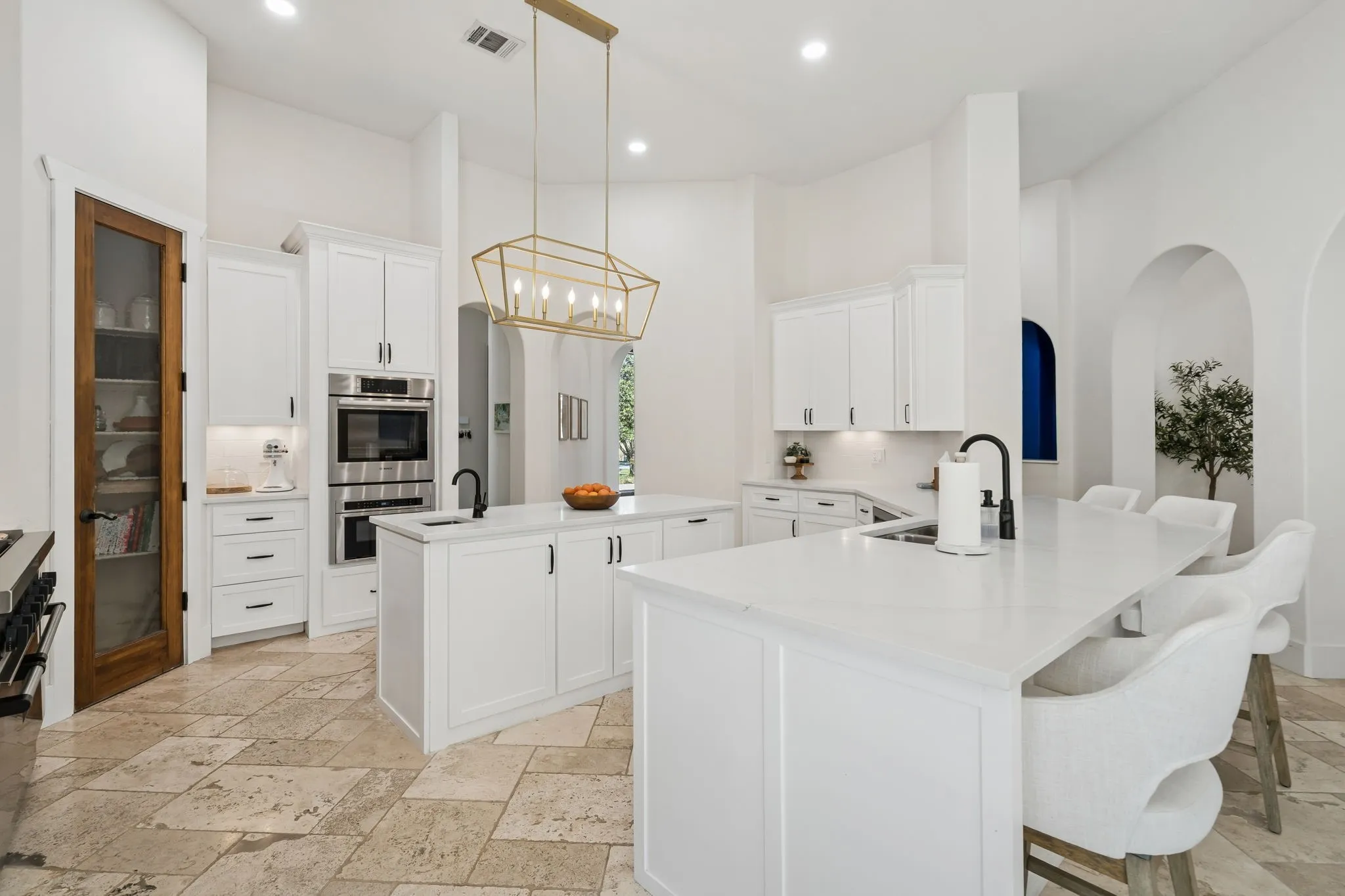 White cabinetry, stone tile flooring, built-in stainless steel appliances, dual kitchen islands with white countertops, and arched wall openings