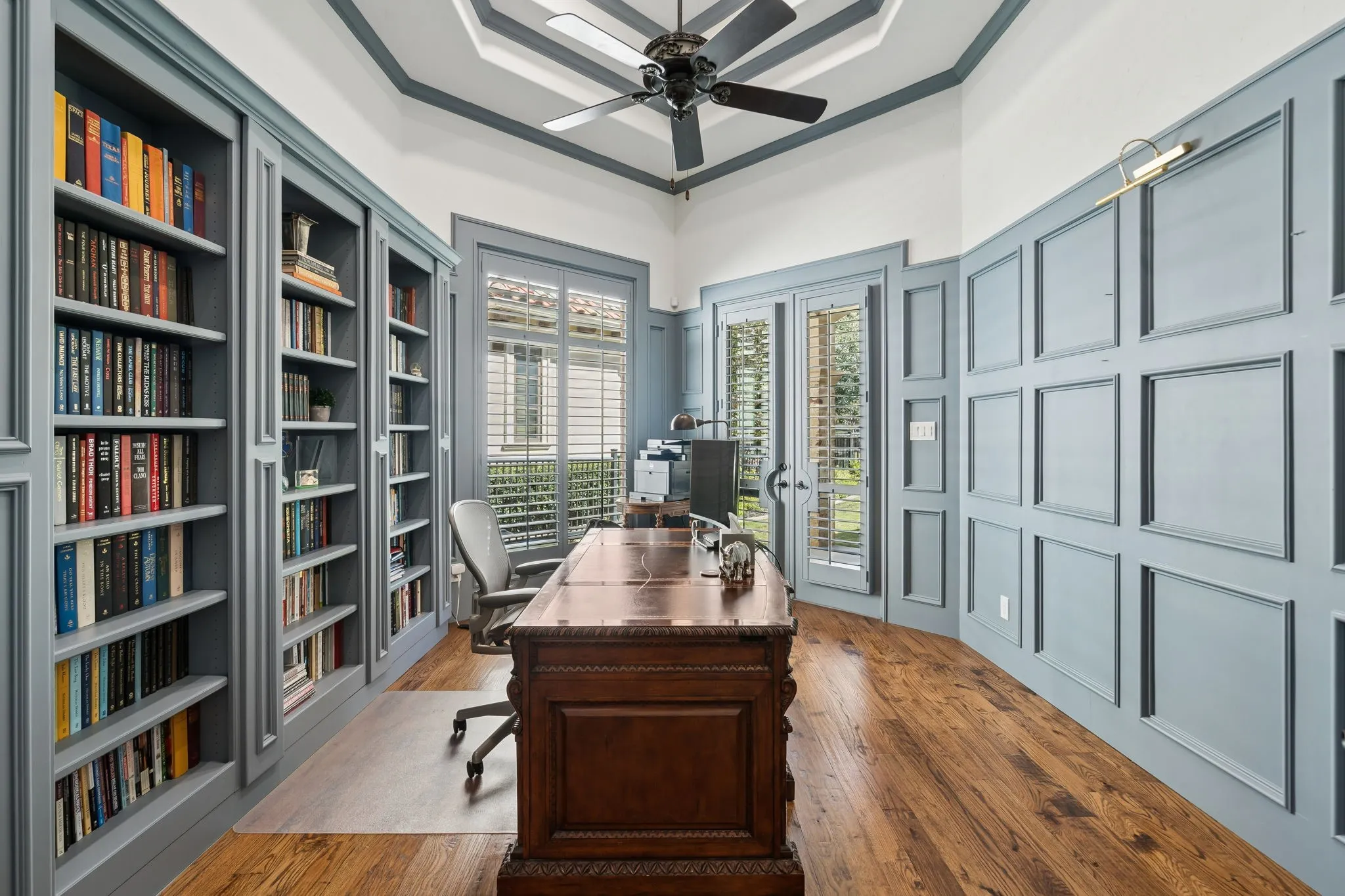 Built-in bookshelves, wood-finish flooring, accent paneling, and a tray ceiling with a ceiling fan