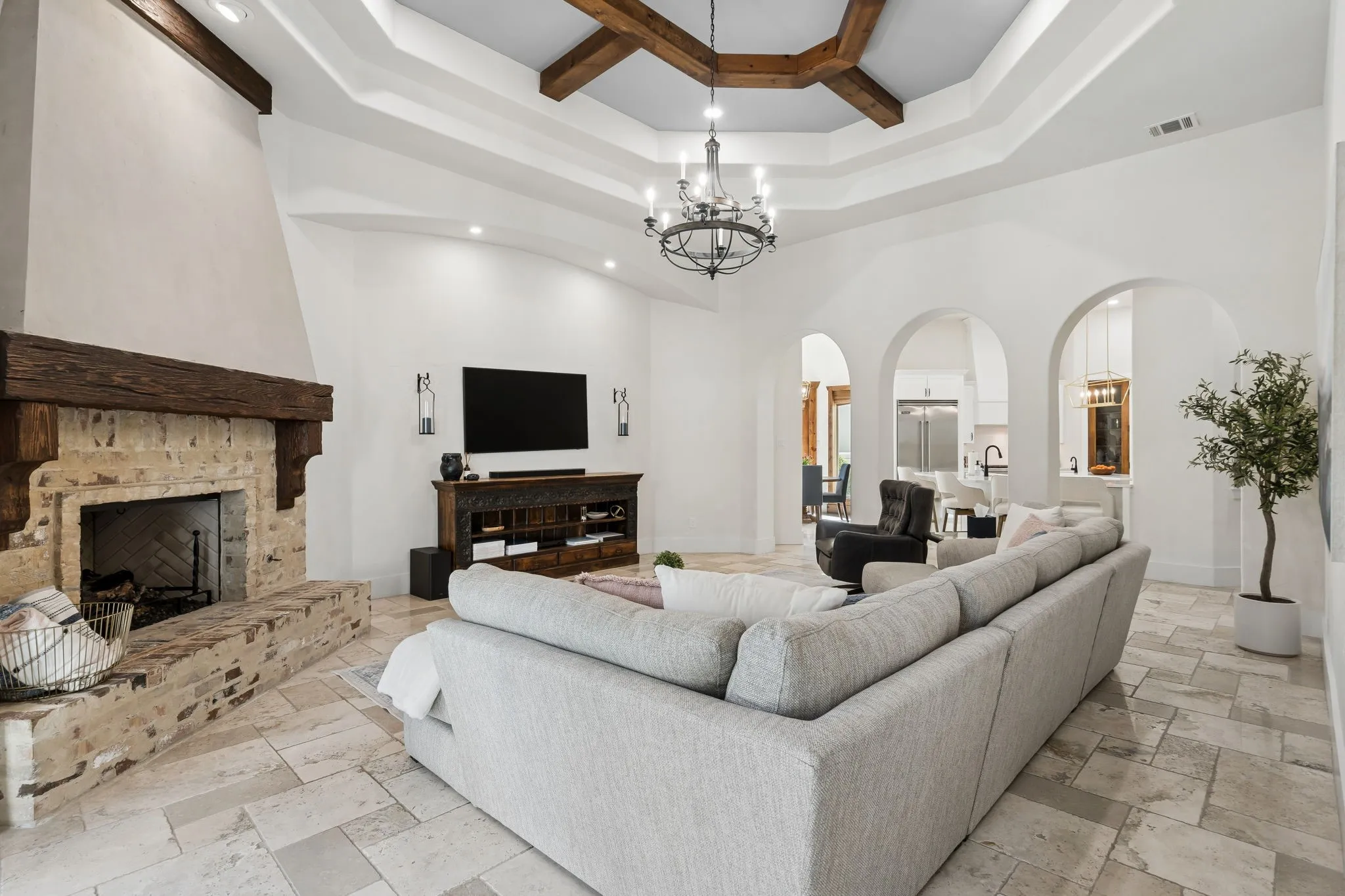 Vaulted ceiling with exposed wood beams, stone fireplace with a substantial wood mantel, arched openings, recessed lighting, and large format tile flooring