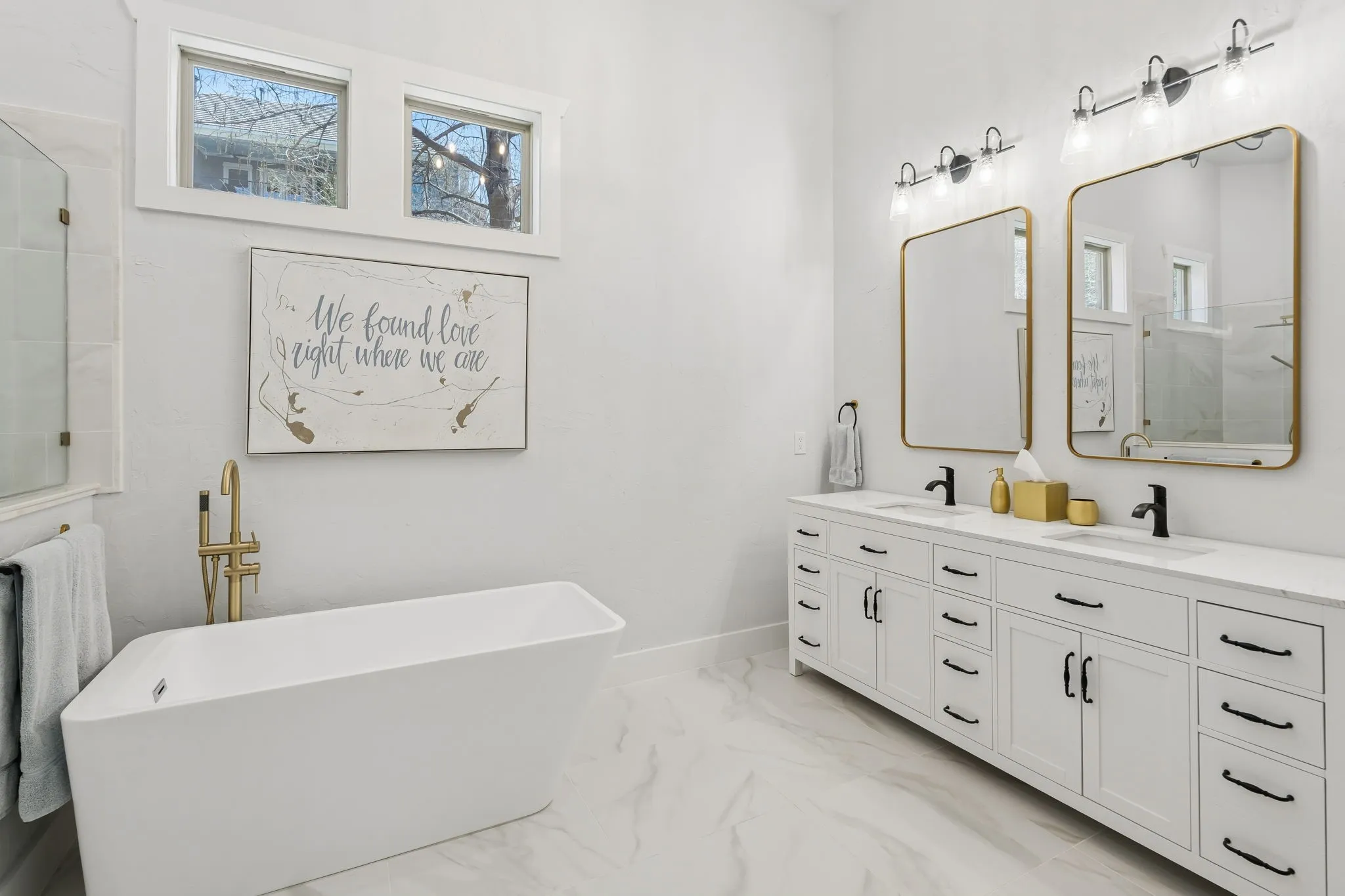 Freestanding soaking tub with a floor-mounted brass faucet, dual vanity with white cabinetry, matte black hardware, and rectangular mirrors, and a glass-enclosed shower