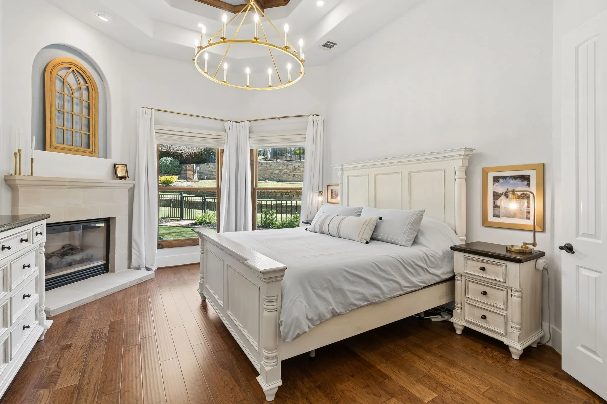 Vaulted ceiling with exposed wood beams, gold-finish chandelier, corner fireplace with stone surround, wood-finish flooring, and multiple windows