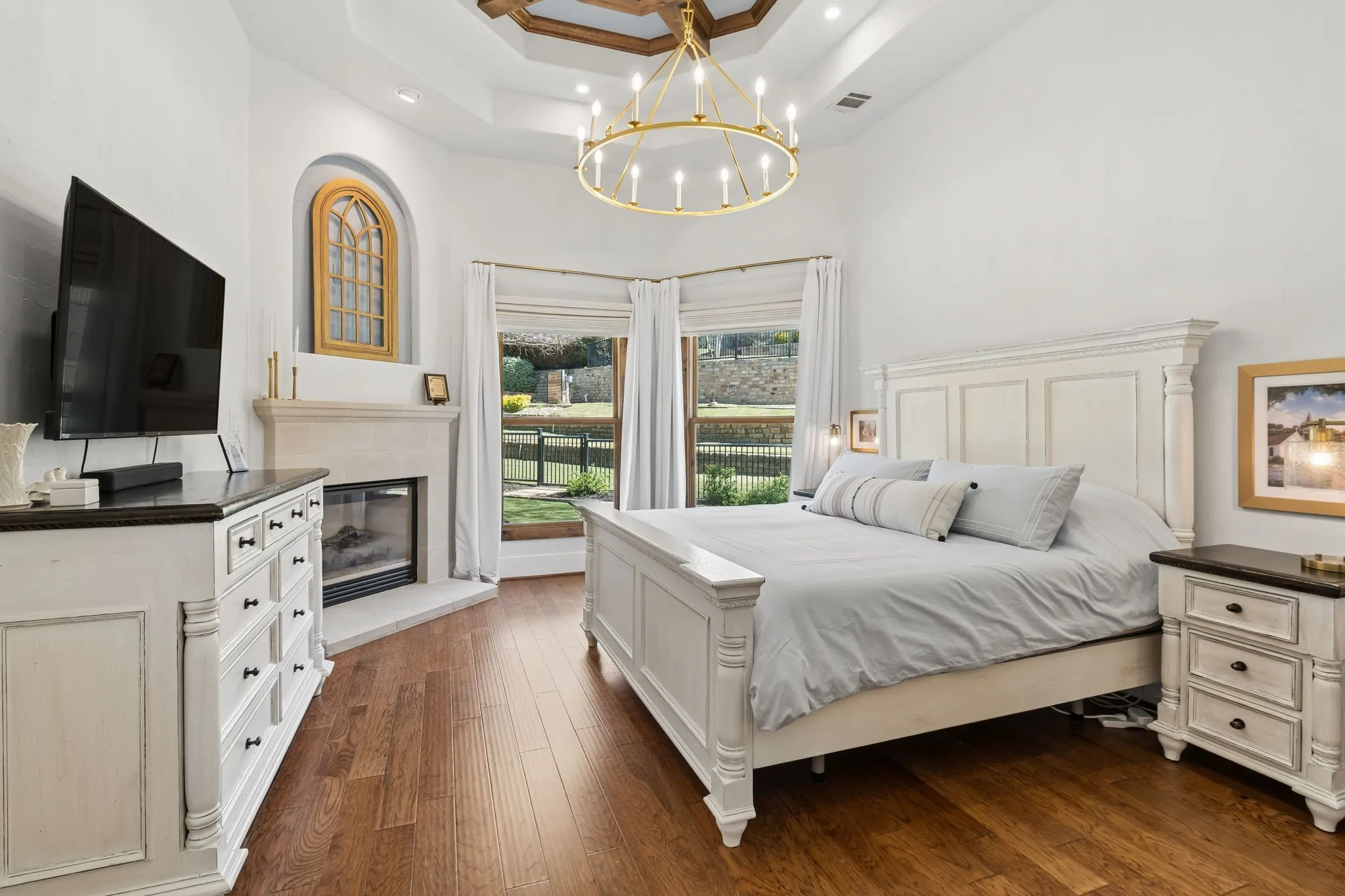 Tray ceiling with wood-finish accents, large ring chandelier, corner fireplace with stone surround, wood-finish flooring, and large windows