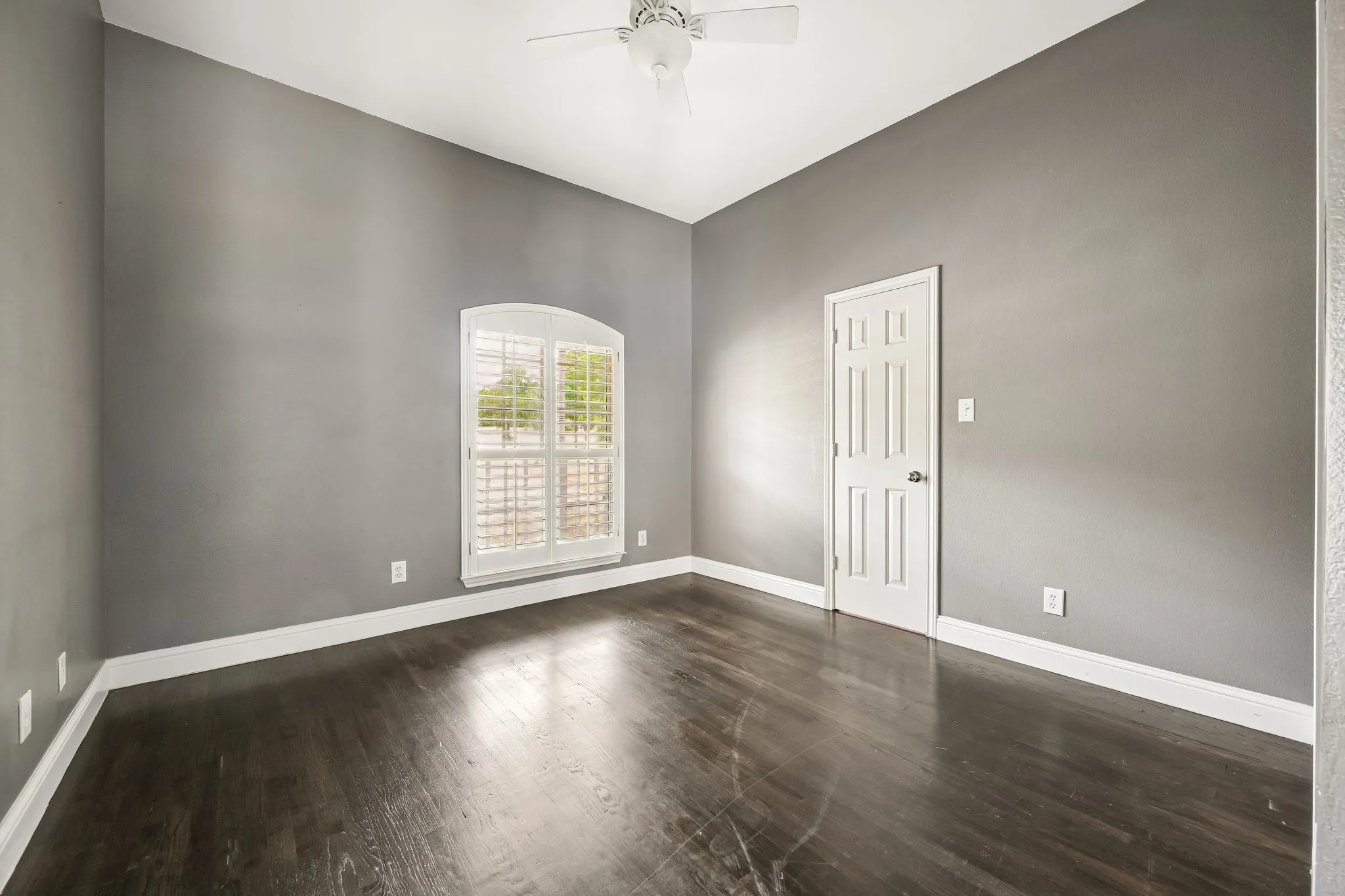 Dark wood-finish flooring, arched window with plantation shutters, ceiling fan, and white trim accents