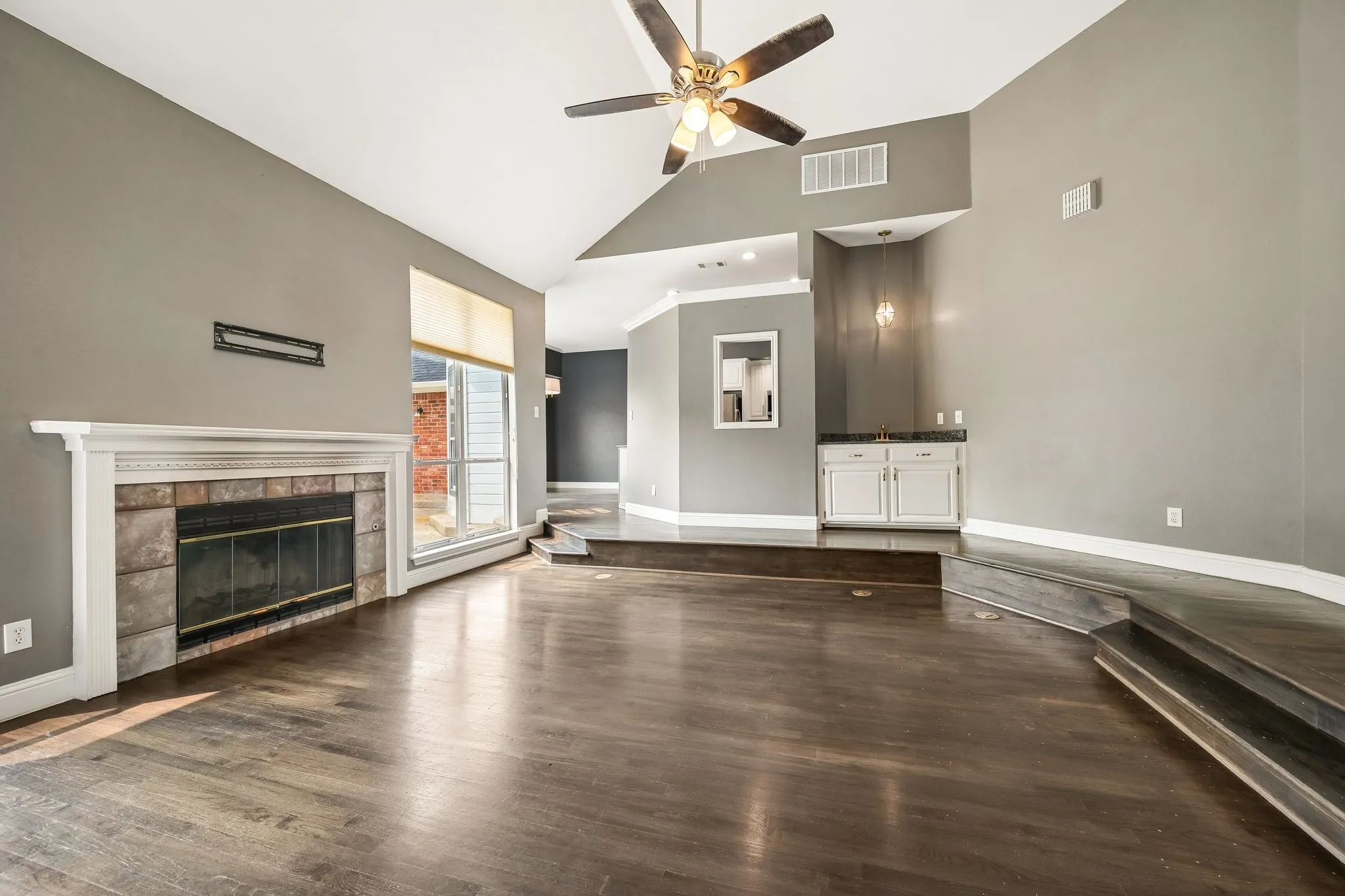 Vaulted ceiling with a ceiling fan, wood-finish flooring, fireplace with stone surround and white mantel, built-in wet bar with white cabinetry and dark countertop, and large window with blinds