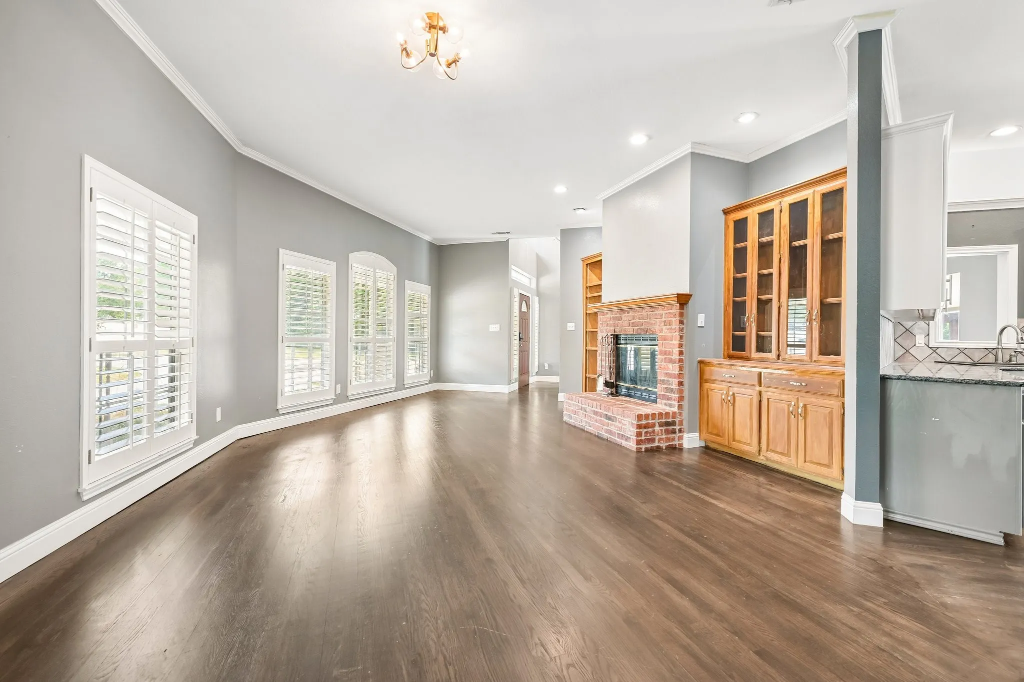 Spacious room featuring wood-finish flooring, a brick fireplace, built-in wooden cabinetry, recessed lighting, and multiple windows with plantation shutters