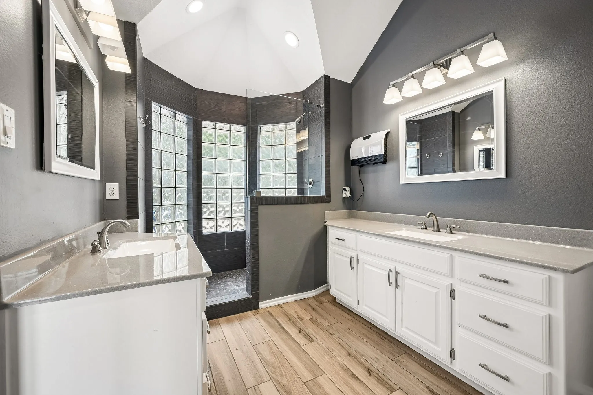 Dual vanity sinks with white cabinetry, wood-finish flooring, and a glass block shower enclosure with dark tile