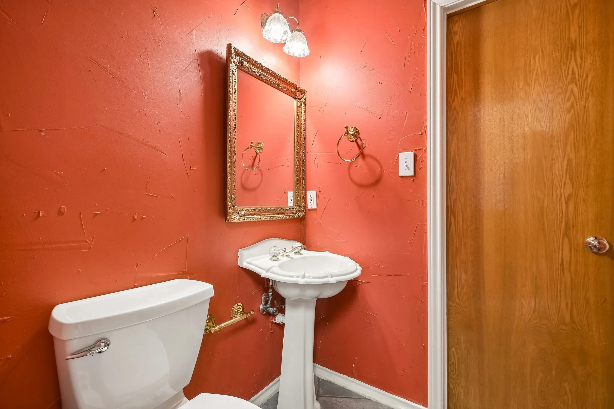 Pedestal sink with a white basin and chrome fixtures, framed mirror with an ornate border, wall-mounted light fixture with two glass shades, and a wood-finish door