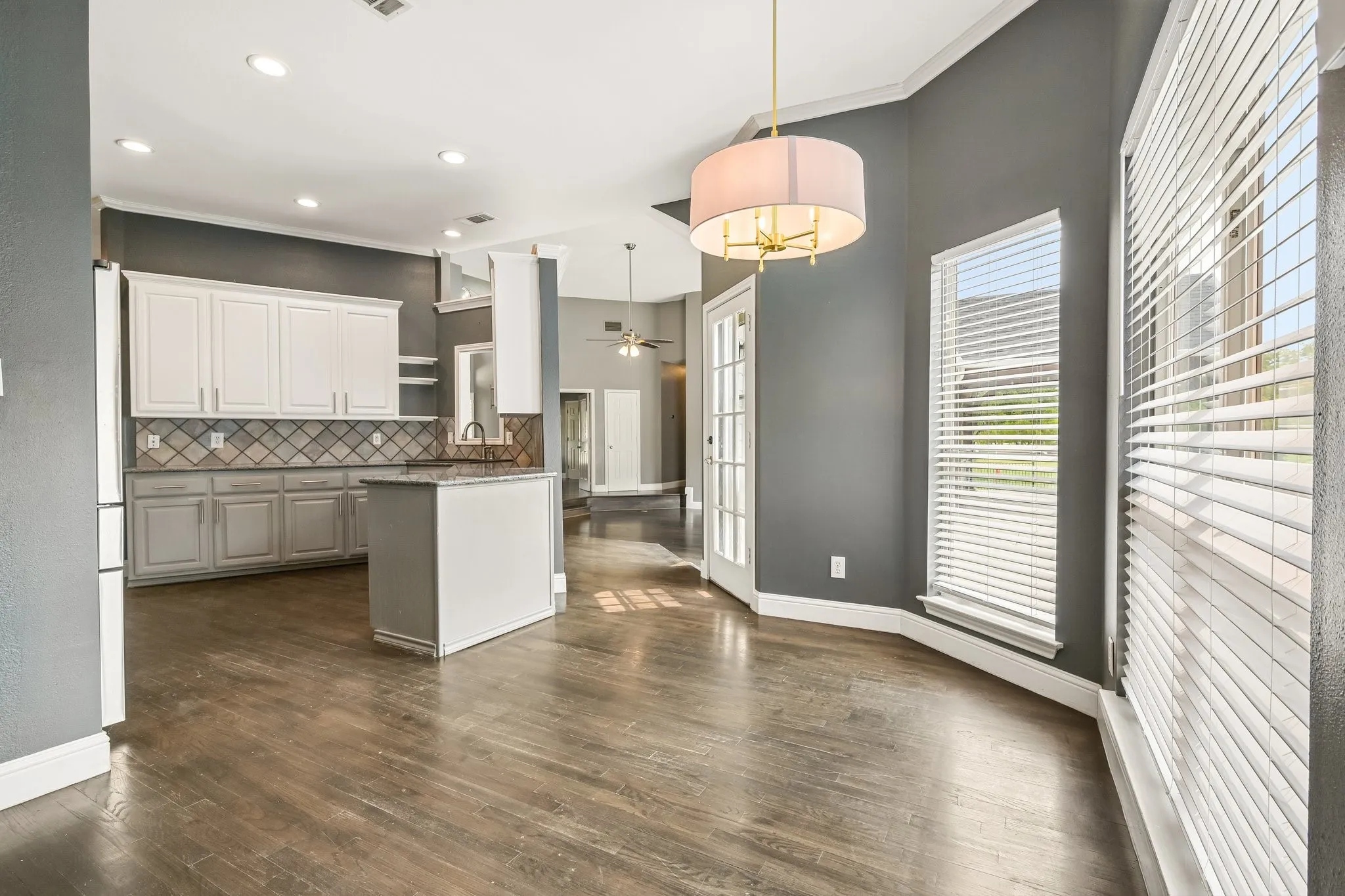 Open-concept kitchen and dining area featuring white cabinetry with decorative tile backsplash, wood-finish flooring throughout, gray accent walls, and a drum pendant light fixture