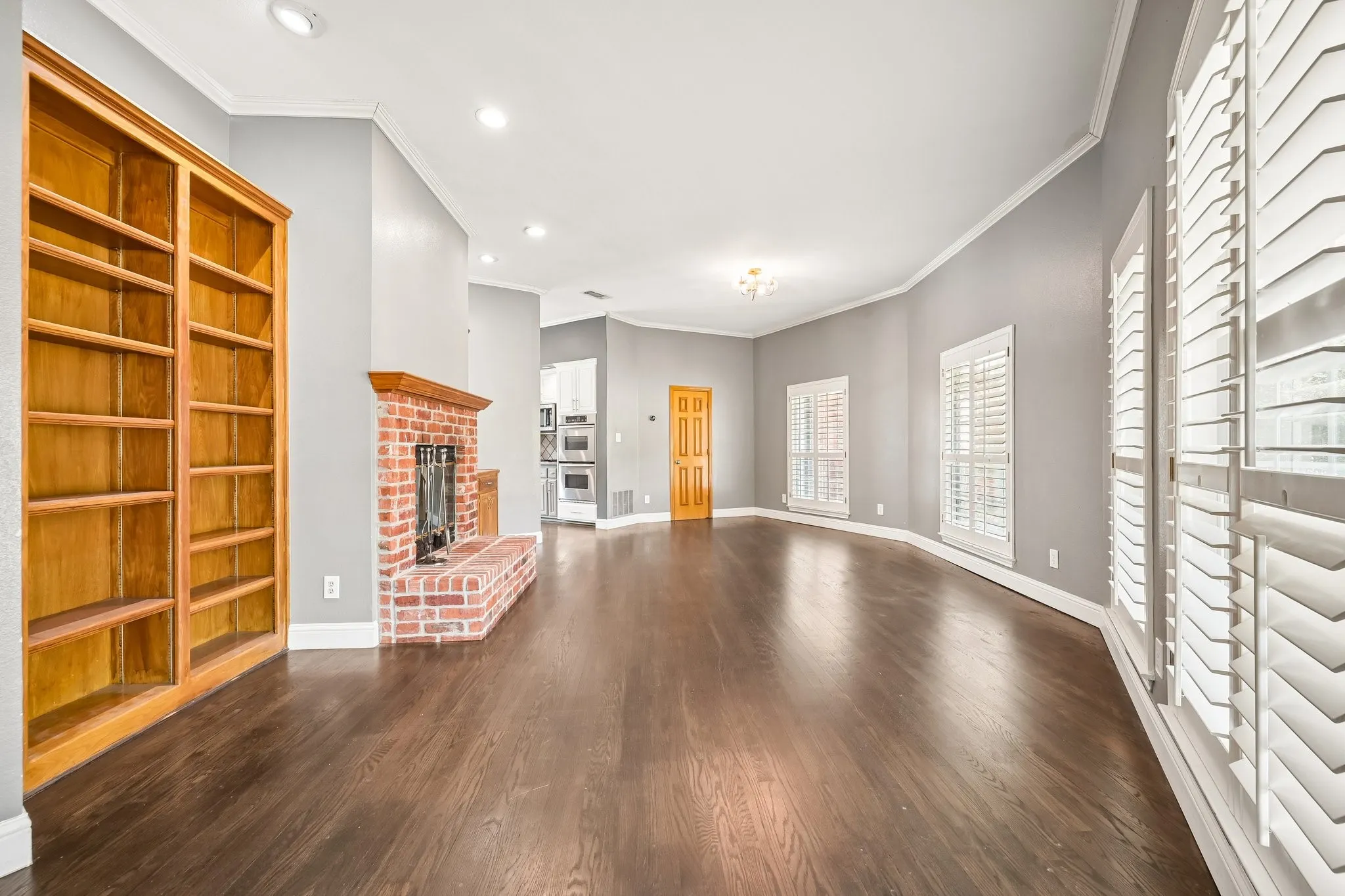 Wood-finish flooring, brick fireplace, built-in wooden shelving, and multiple windows with plantation shutters