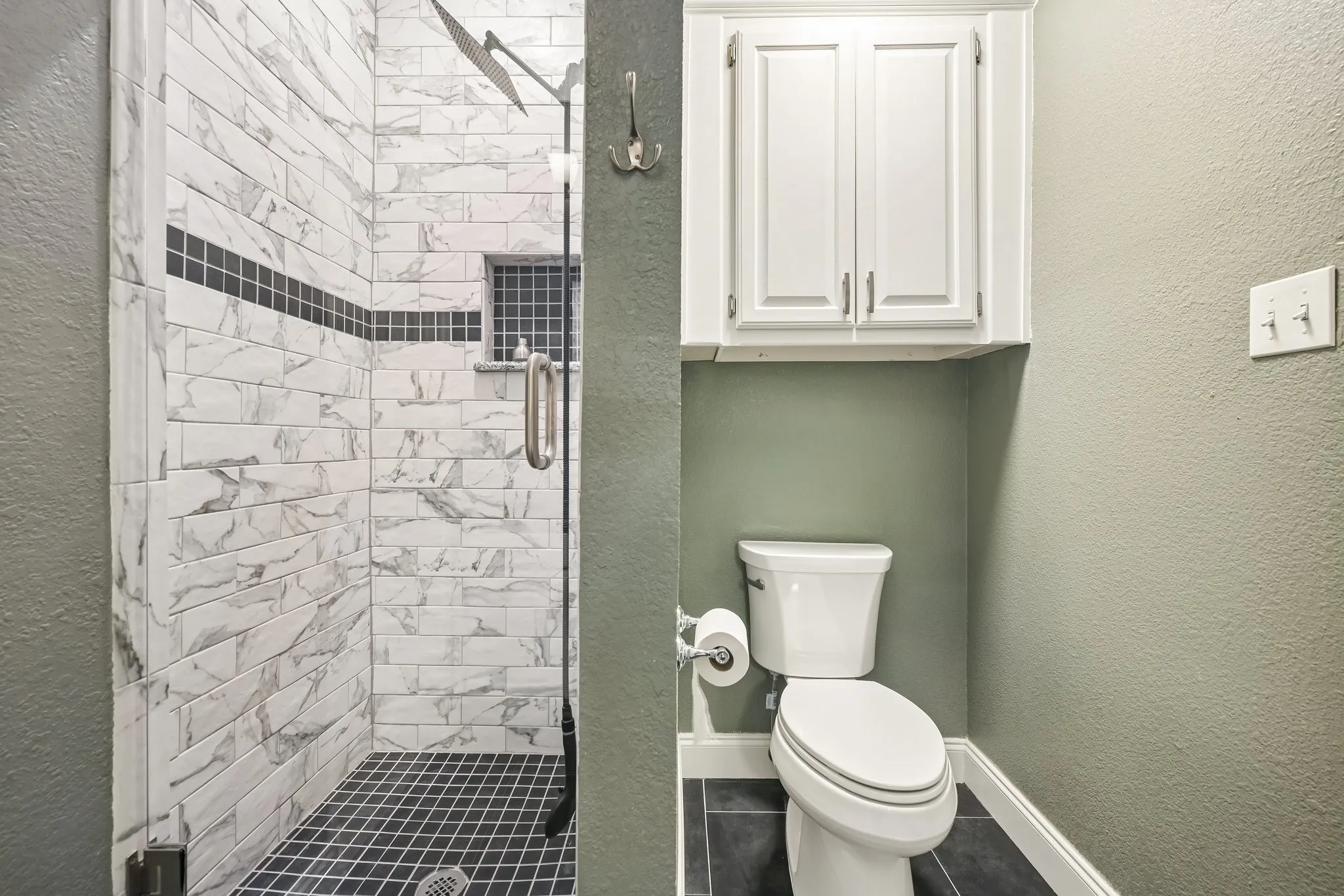 Spa-inspired bathroom featuring marble-look subway tile shower surround with black accent stripe, black hexagonal floor tile, frameless glass shower enclosure, white porcelain toilet with elongated bowl, and white cabinetry with brushed nickel hardware above