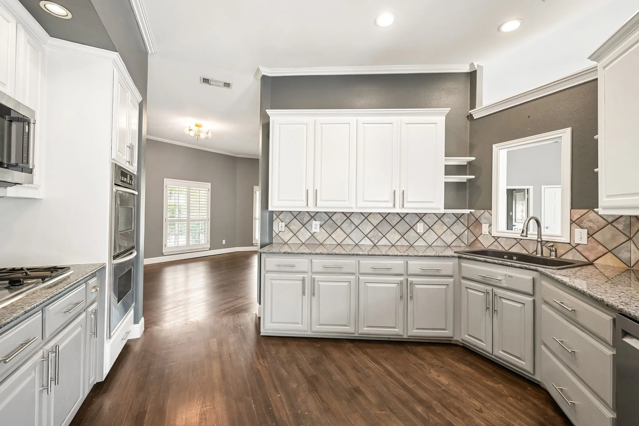 Chef's kitchen featuring white cabinetry with brushed nickel hardware, stainless steel appliances, geometric tile backsplash, granite countertops, and wood-finish flooring throughout the open layout