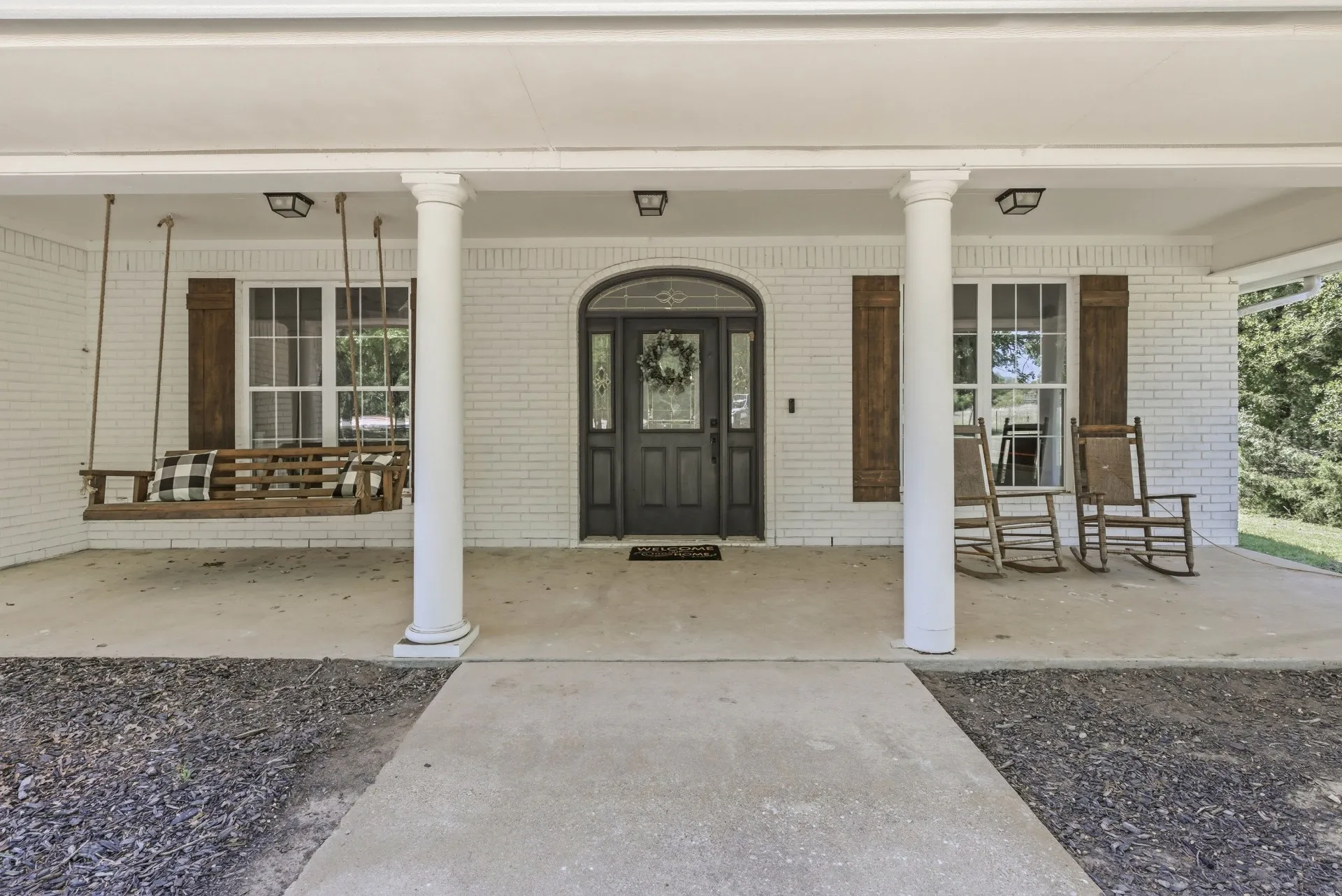Expansive covered porch featuring a painted brick exterior, multiple white columns, and a dark wood-finish entry door with an arched transom