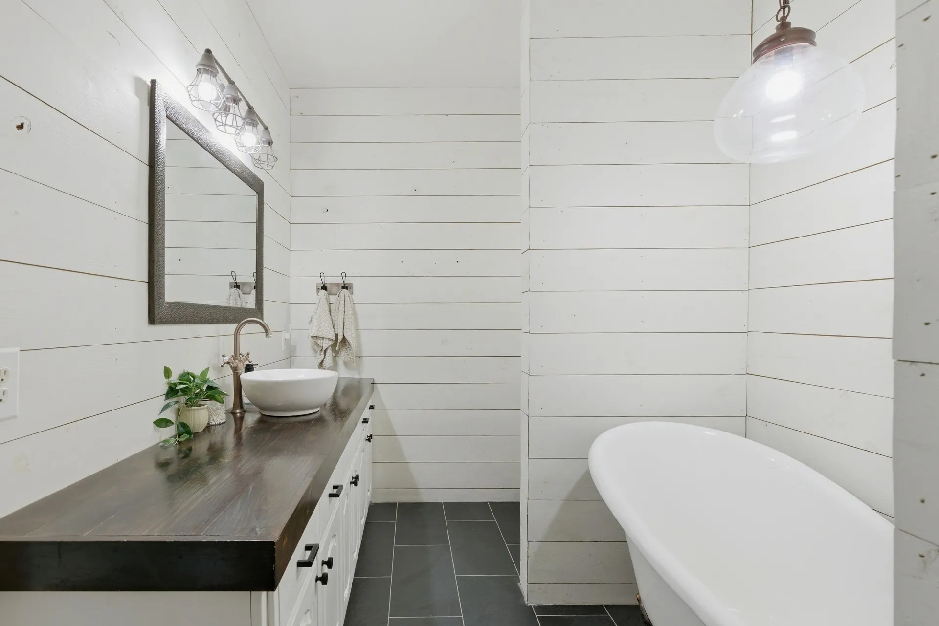 White shiplap walls with a freestanding bathtub, dark wood-finish countertop, and a vessel sink with a bronze-finish faucet