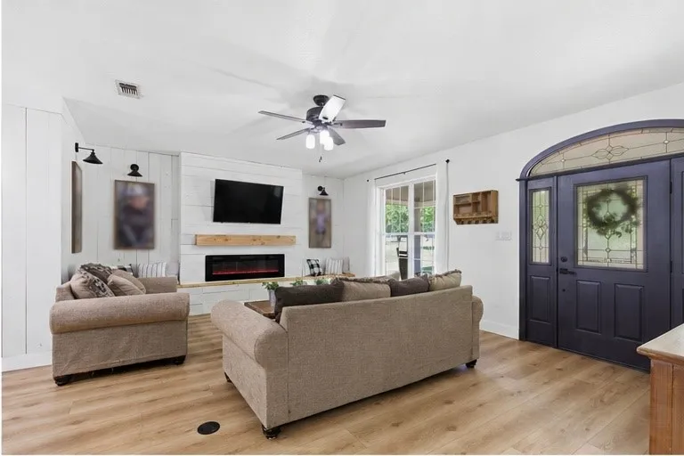 Wood-finish flooring, ceiling fan, white shiplap fireplace surround with wood mantel, built-in electric fireplace, and an arched entry door with decorative glass