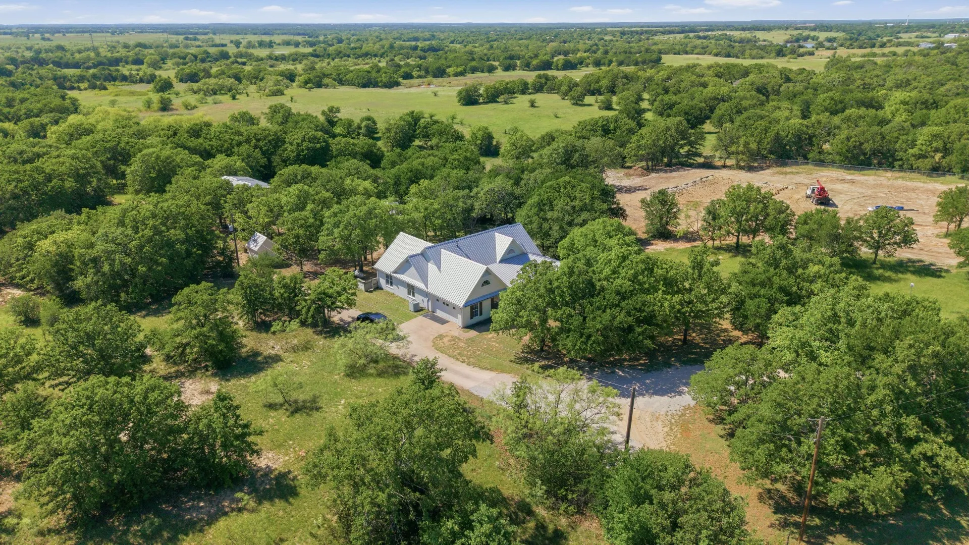 Expansive property featuring a residence with a light-colored exterior and a metal roof, surrounded by numerous mature trees
