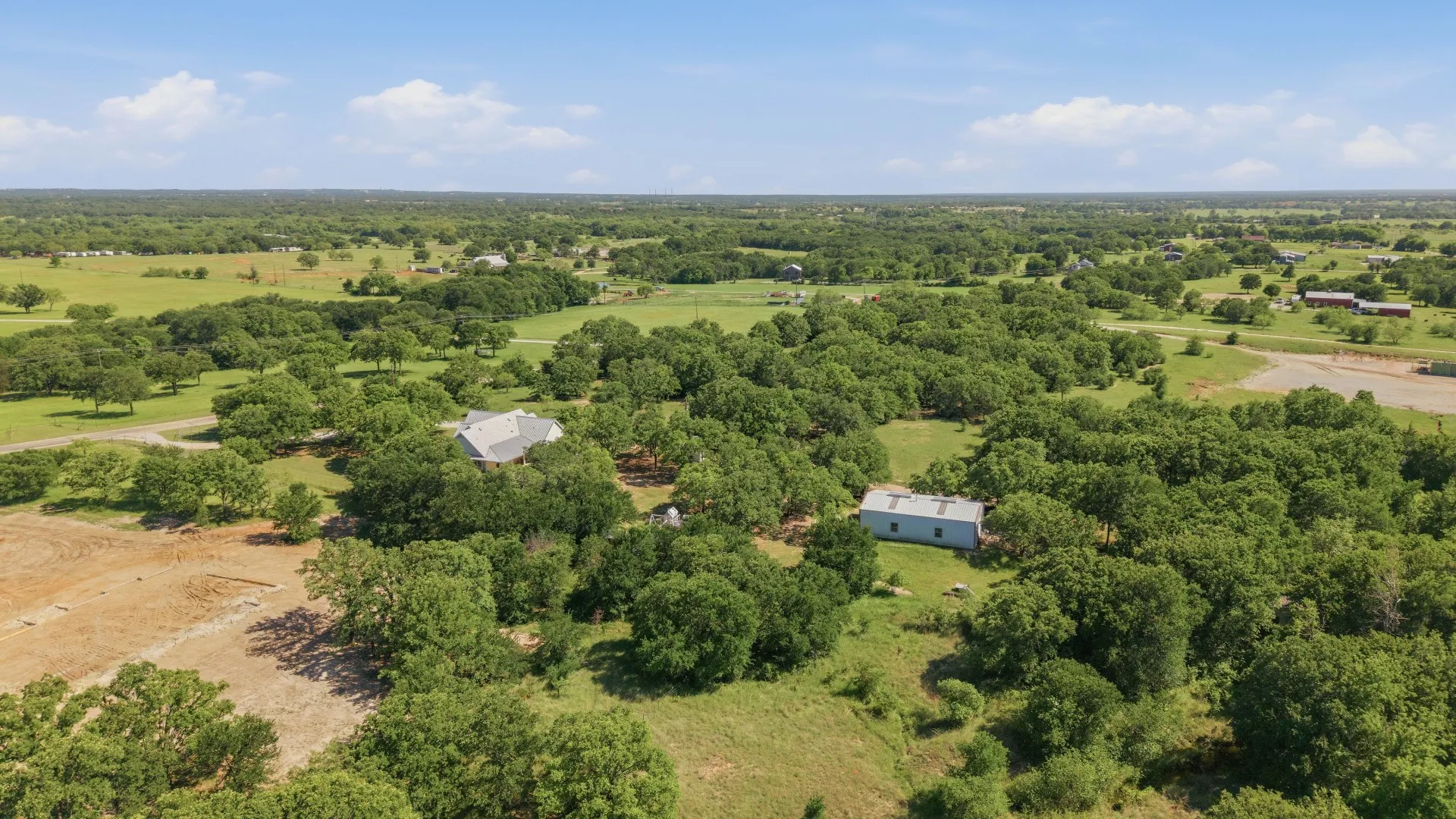 Expansive acreage with mature trees, a single-story residence, and an auxiliary building, all set under a clear sky