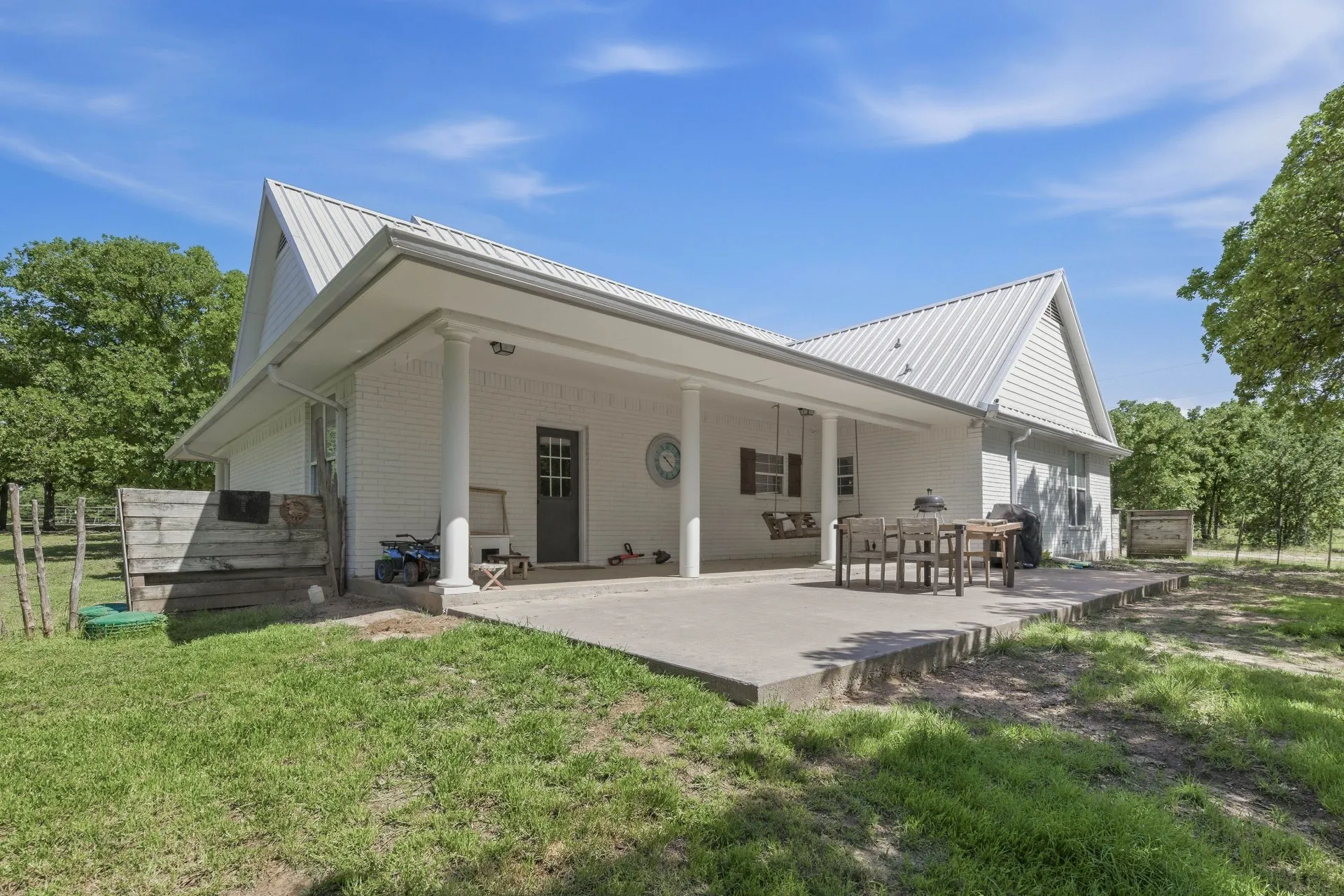White brick exterior with a metal roof, featuring a covered concrete patio supported by columns, and a partially fenced perimeter