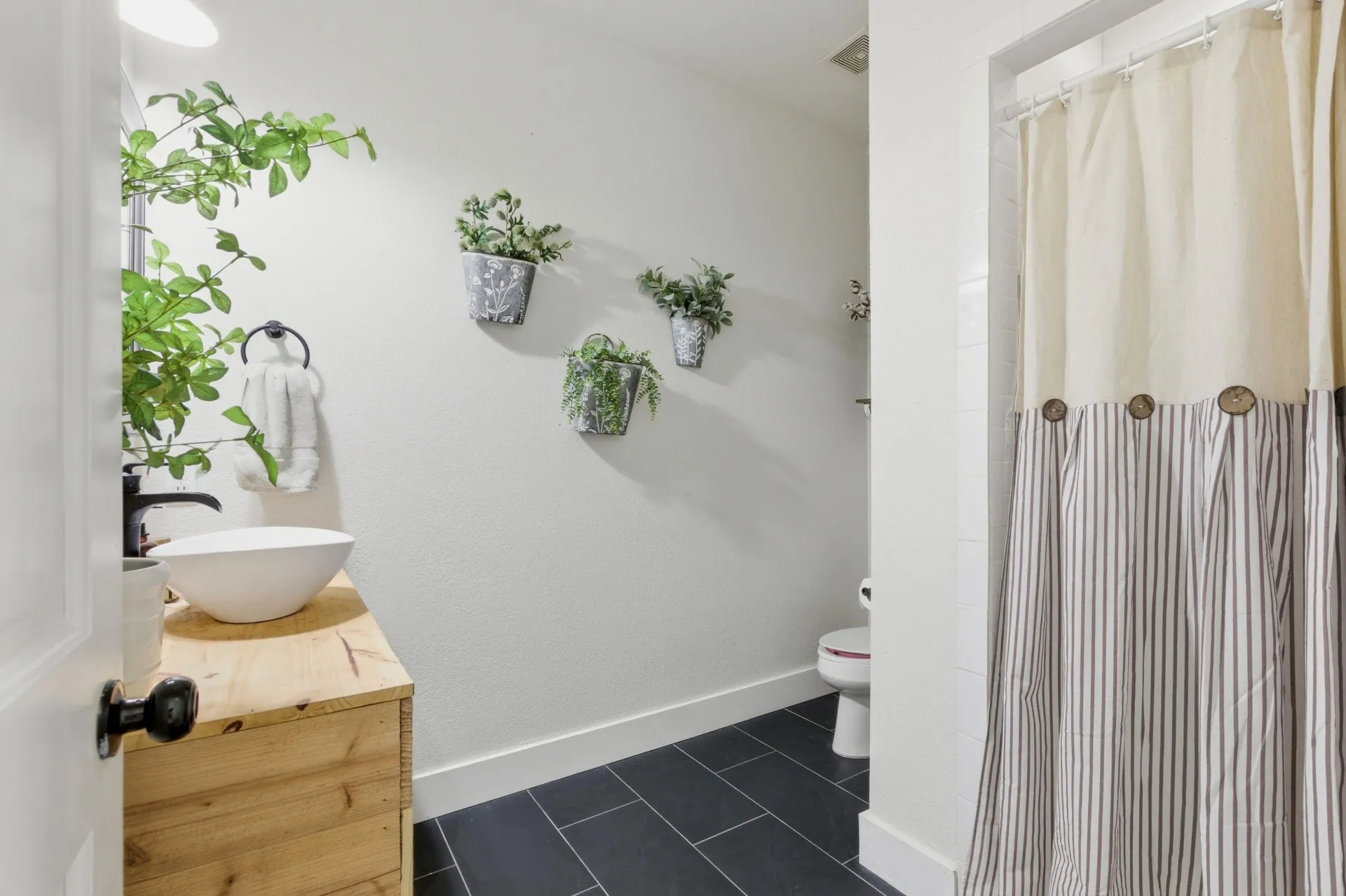 White vessel sink with matte black faucet, natural wood vanity, dark rectangular floor tiles, and a built-in toilet