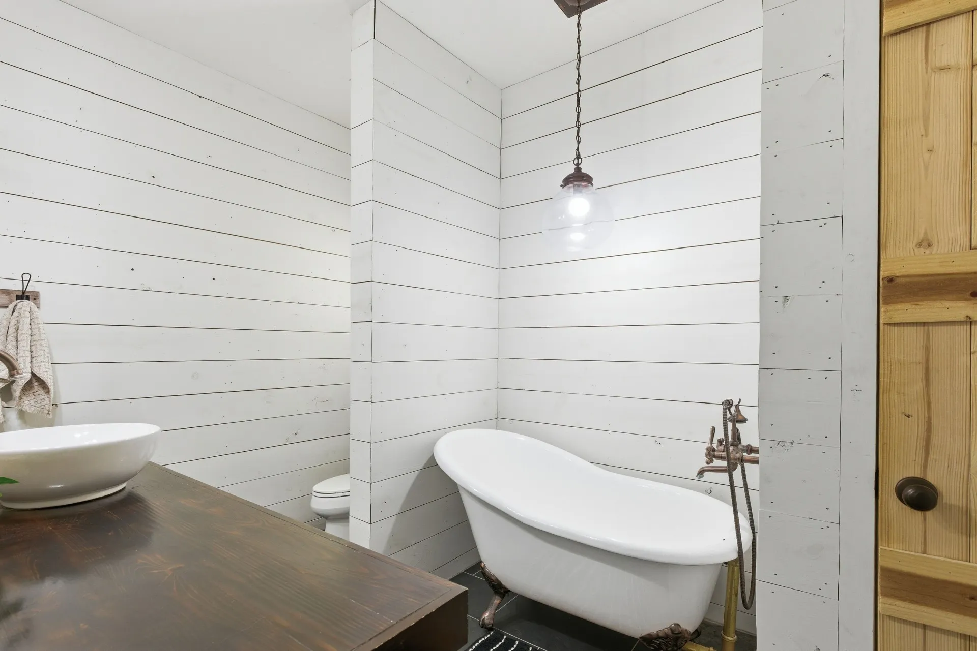 White shiplap walls, dark wood-finish vanity, clawfoot tub with rustic fixtures, vessel sink, and a natural wood-panel door