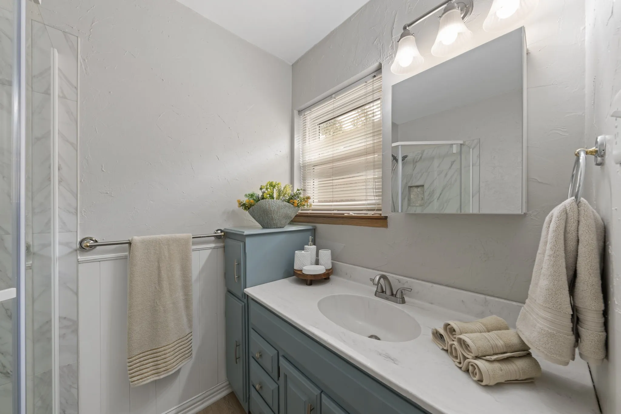 Shower enclosure with glass panel, white paneled wainscoting, window with wood trim, vanity with blue cabinetry, and white countertop with integrated sink