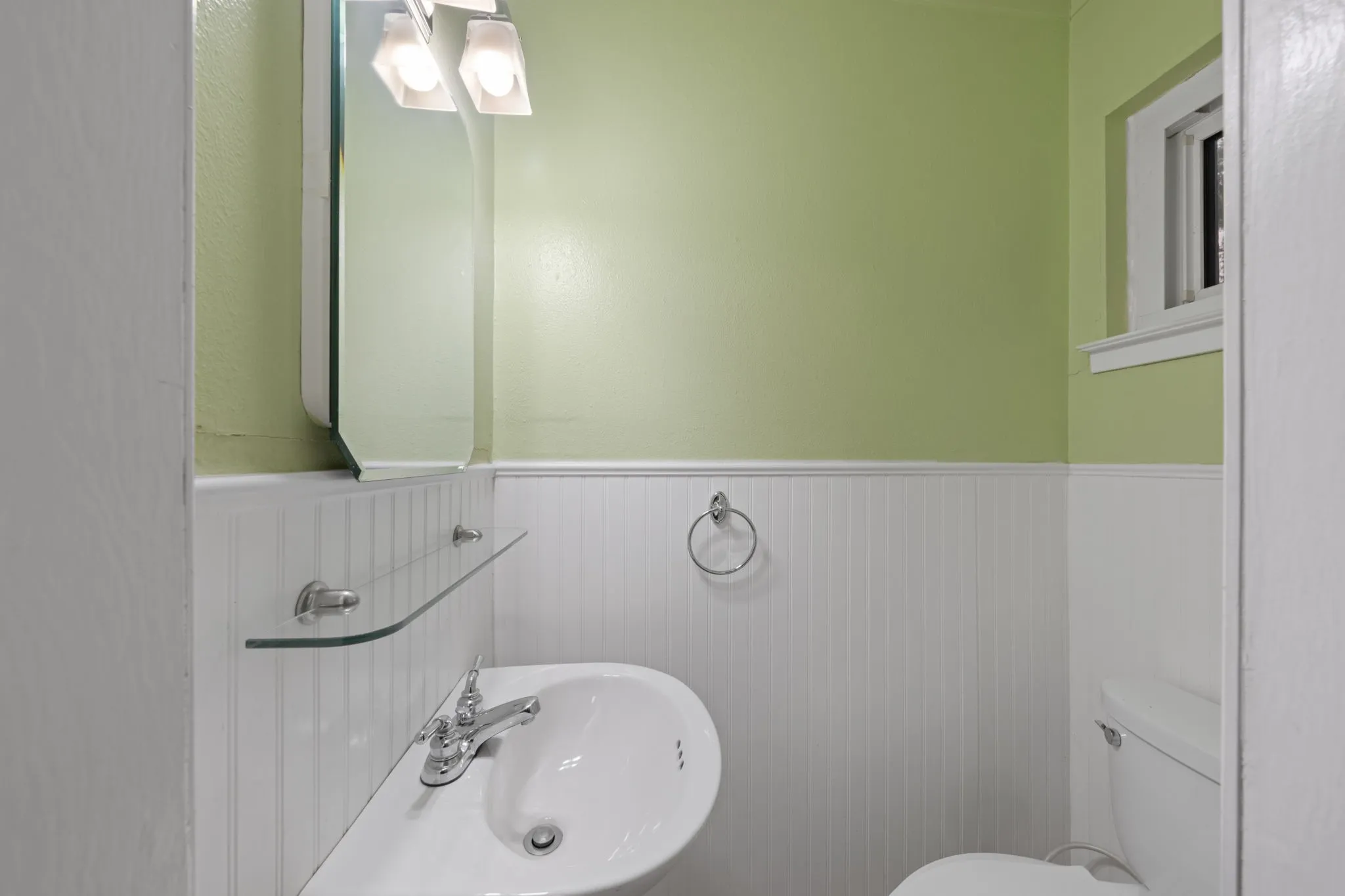 White paneled wainscoting with a pale green wall, corner sink with chrome faucet, wall-mounted mirror, and overhead light fixture