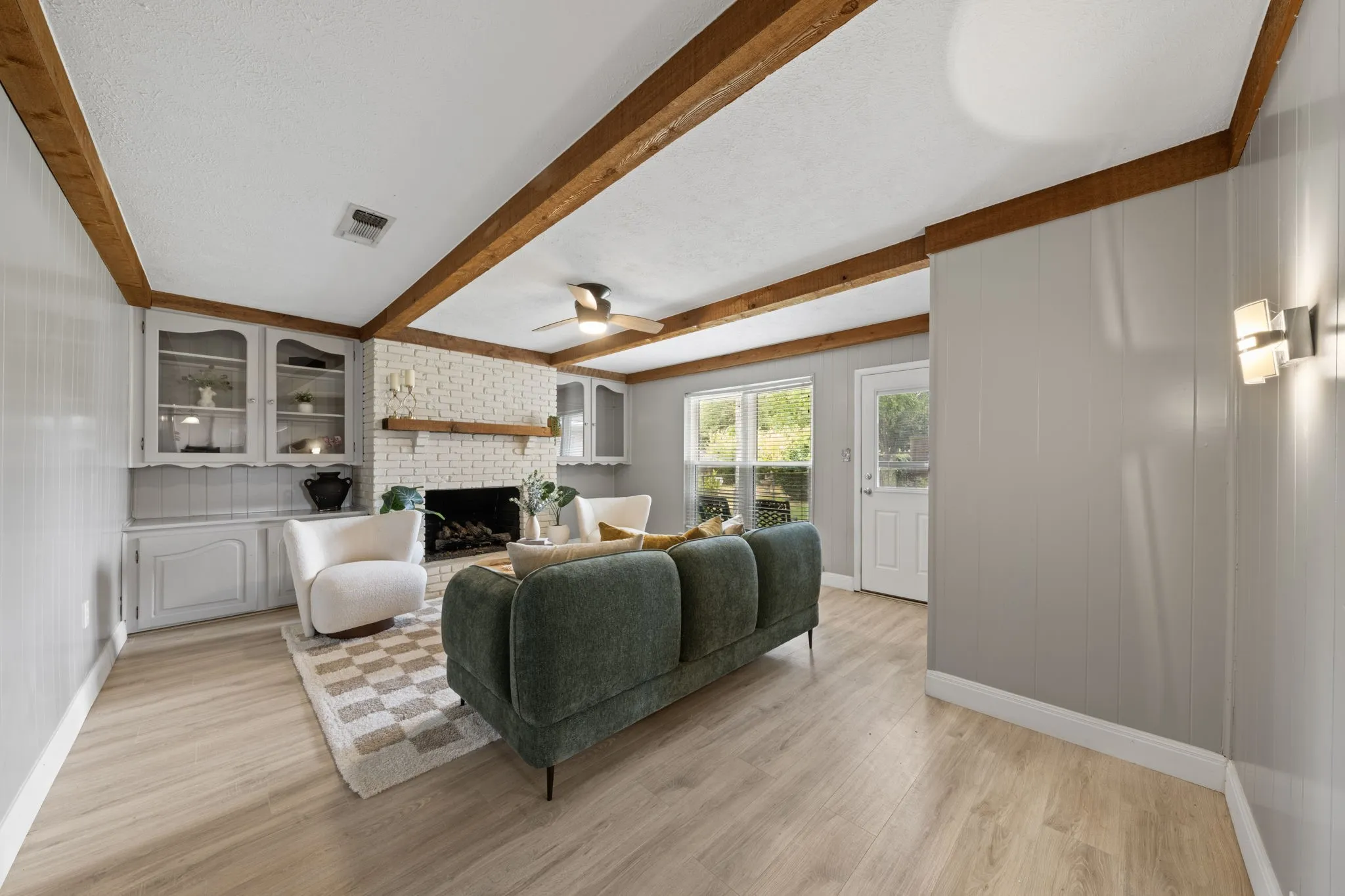 Exposed wood beams, wood-finish flooring, built-in cabinetry with glass doors, white brick fireplace, and modern wall sconces