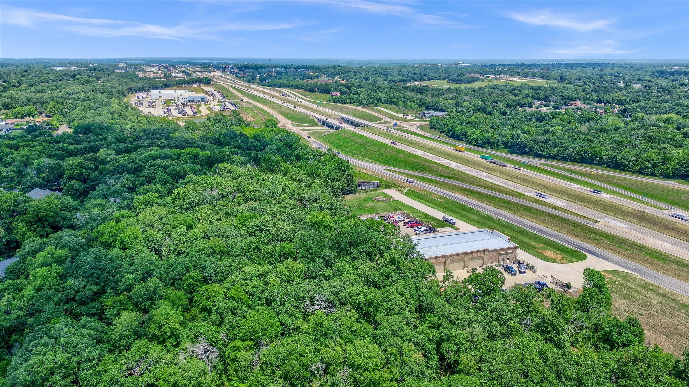 Expansive tree coverage, adjacent multi-lane highway, commercial building with paved lot, and surrounding developed areas