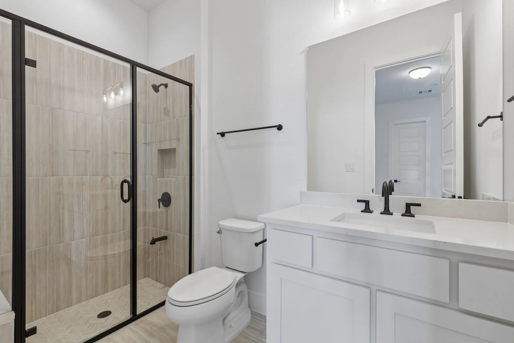 Frameless glass shower enclosure with matte black fixtures, integrated shower niche, a single vanity with a white countertop, and a large wall-mounted mirror