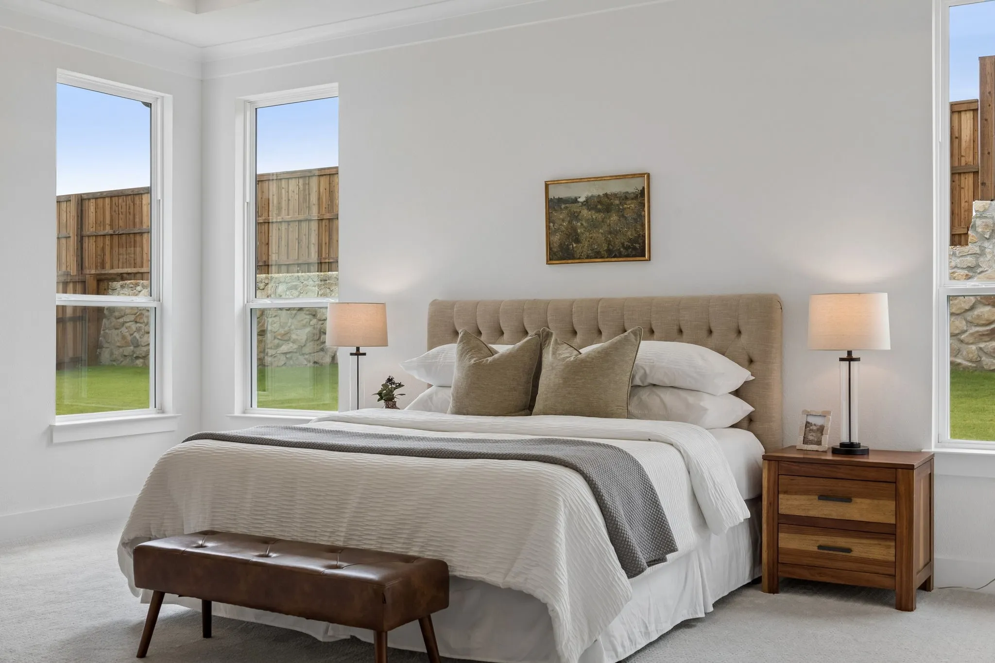 White walls, multiple windows with white trim, light-colored carpeting, and a built-in upholstered headboard