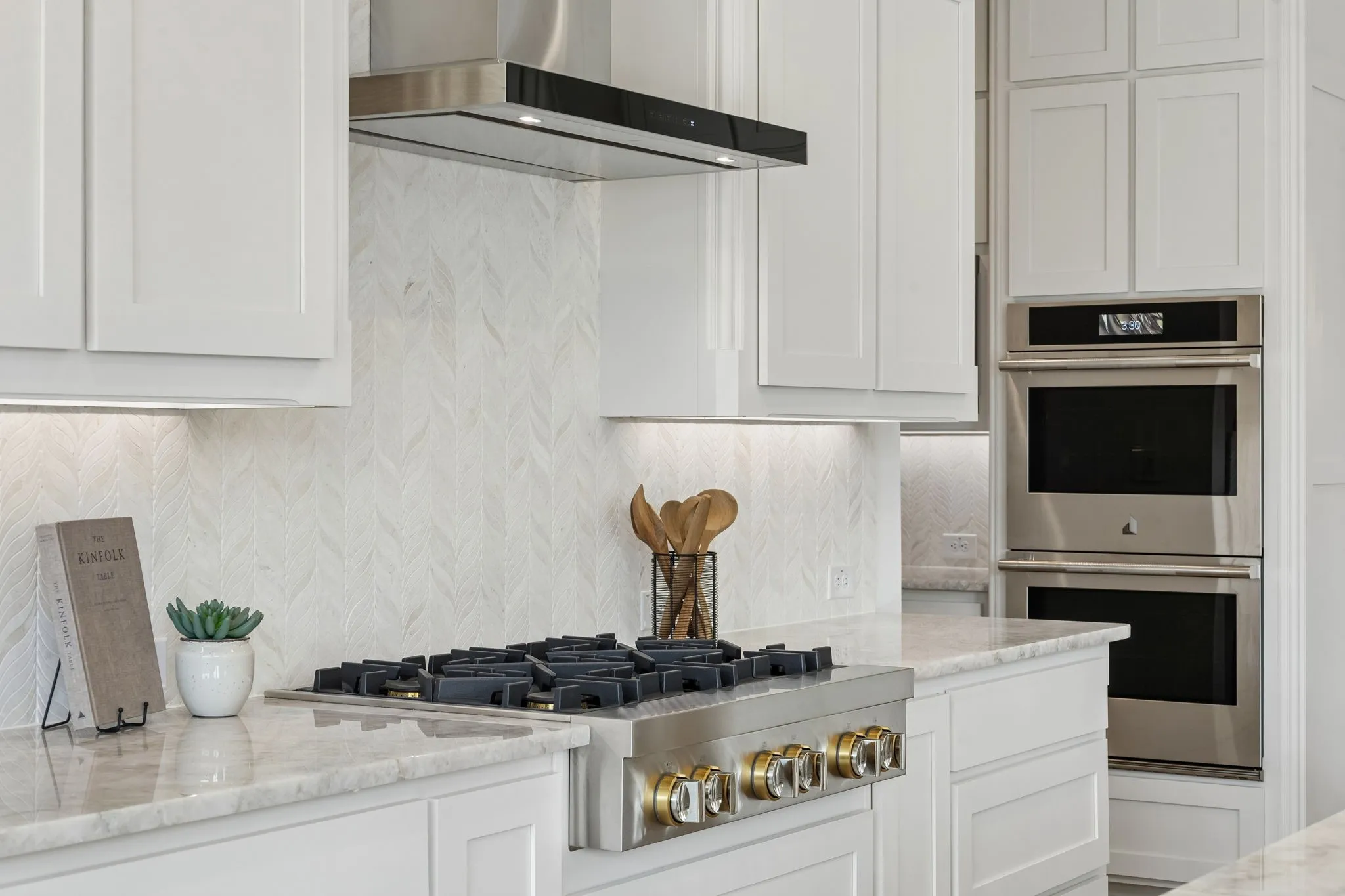 White shaker cabinetry, stainless steel range hood, herringbone pattern backsplash, built-in double ovens, and stone countertops