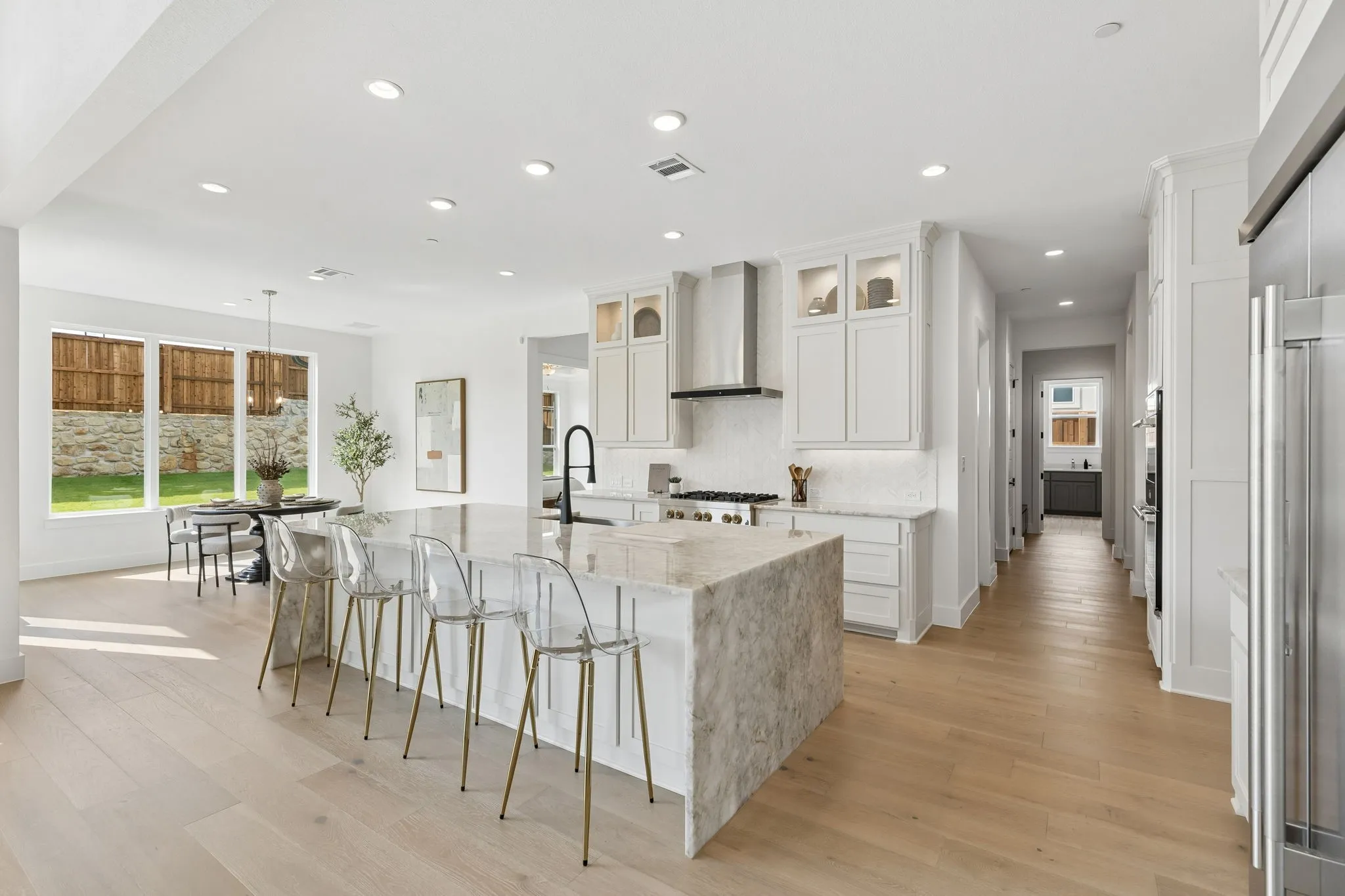 Gourmet kitchen with a large waterfall island, stainless steel range hood, built-in ovens, and light wood-finish flooring