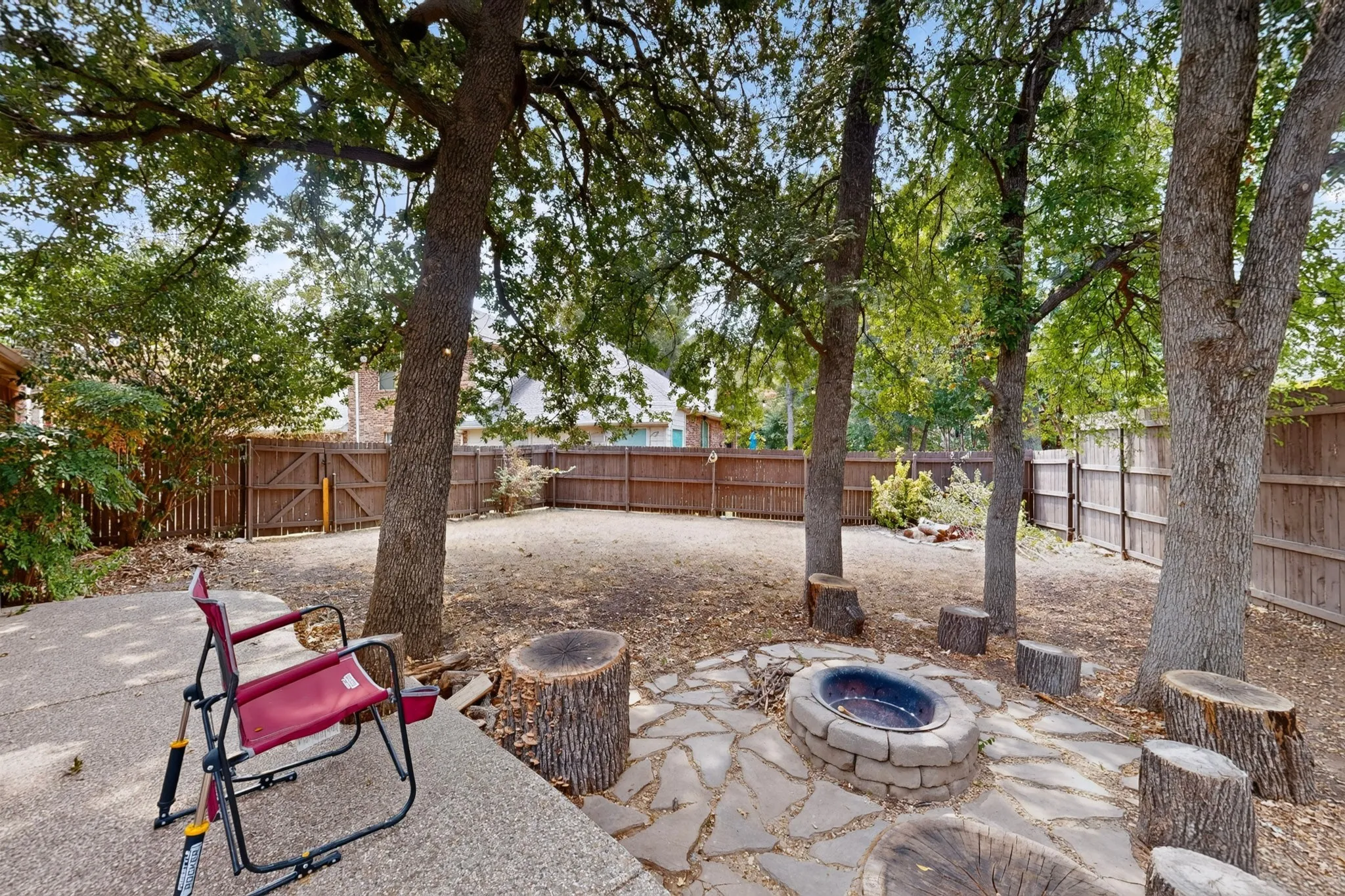 Expansive backyard featuring mature trees, a wood privacy fence, a concrete patio area, and a stone-lined fire pit with flagstone paving