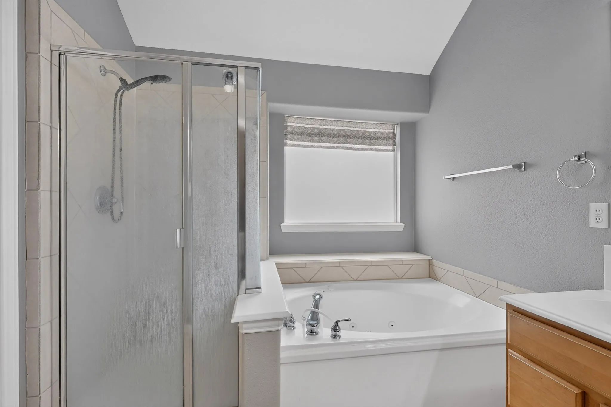 Soaking tub with jetted features, glass-enclosed shower, light wood-finish vanity with countertop, and a window with a roller shade