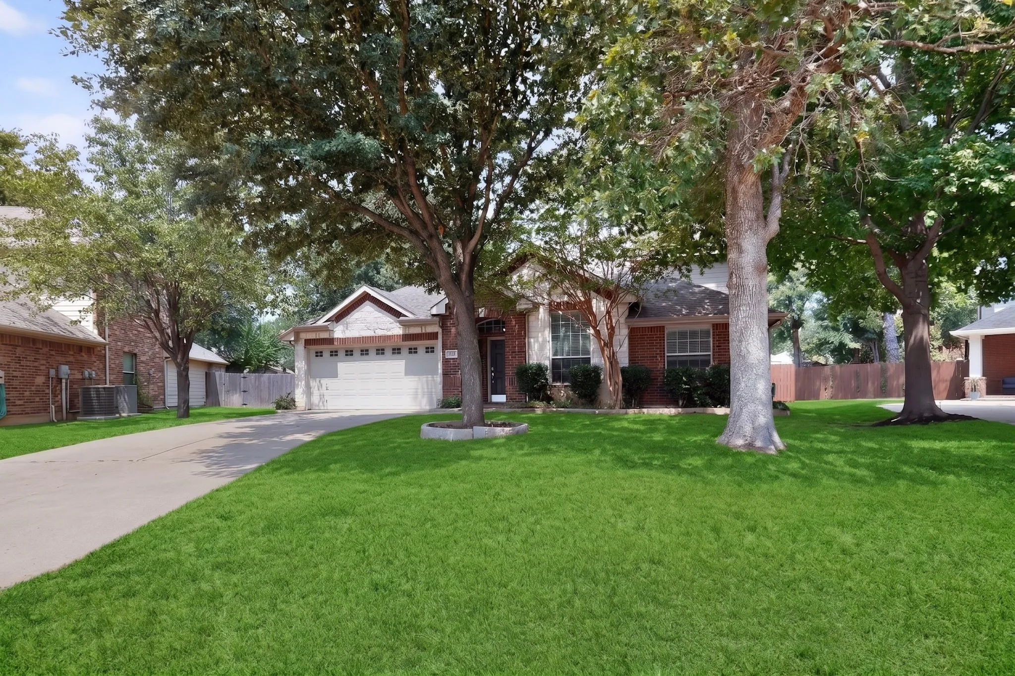 Brick exterior with a gabled roofline, attached garage, mature trees, and a concrete driveway