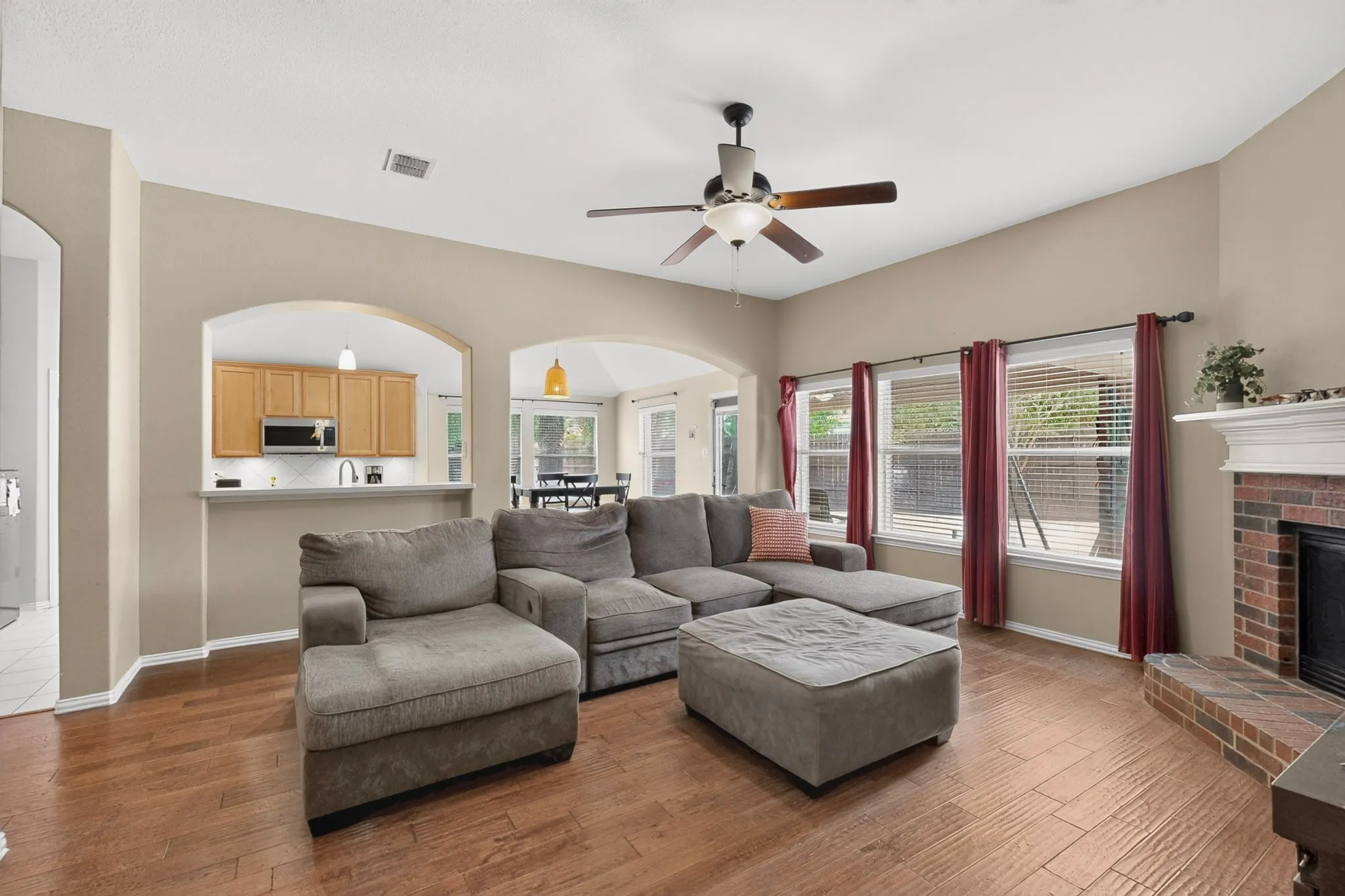 Spacious living area featuring wood-finish flooring, a brick fireplace, a ceiling fan, and multiple windows