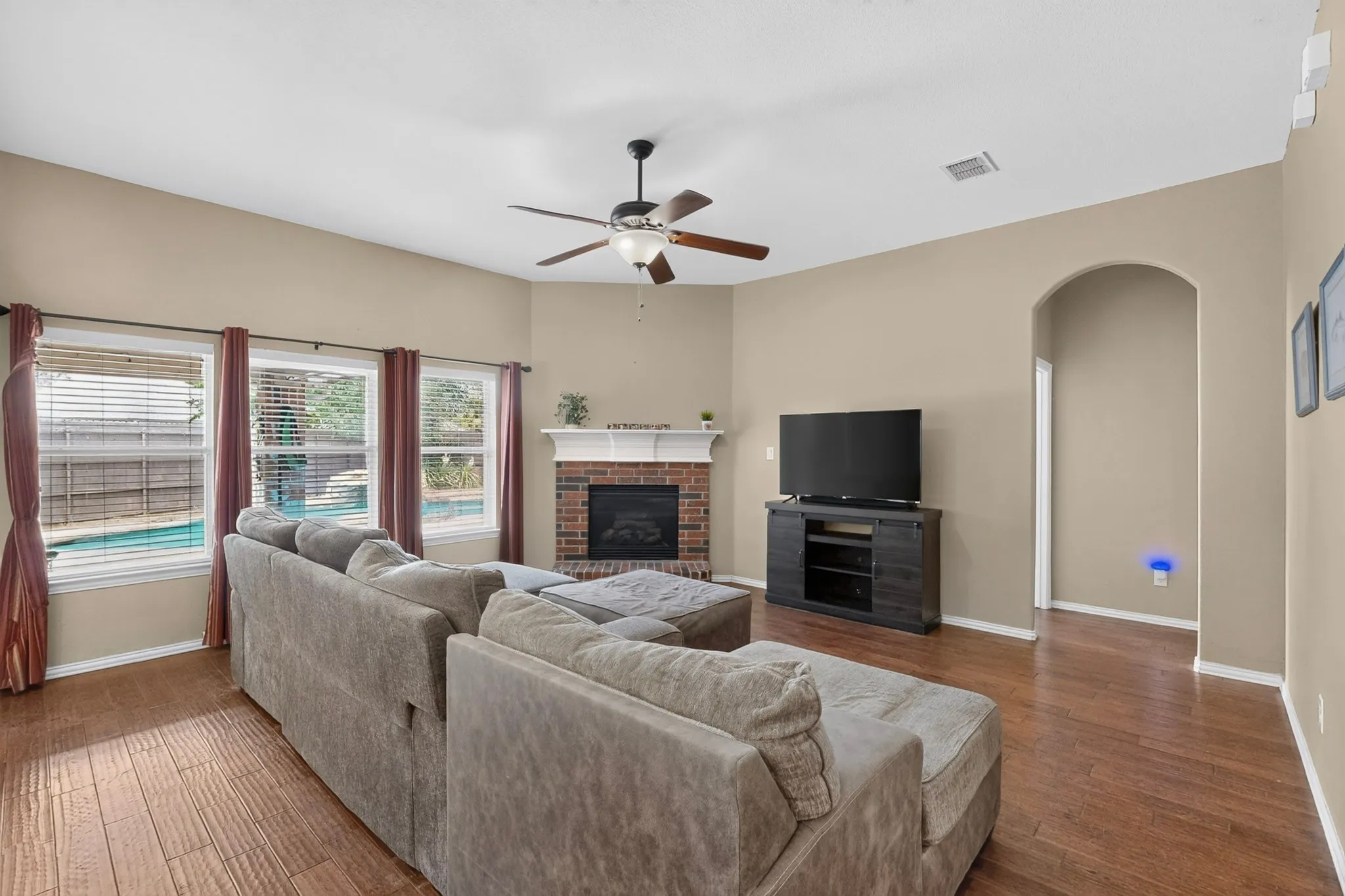 Living area with wood-finish flooring, a corner brick fireplace with a white mantle, an arched doorway, and multiple windows offering external views