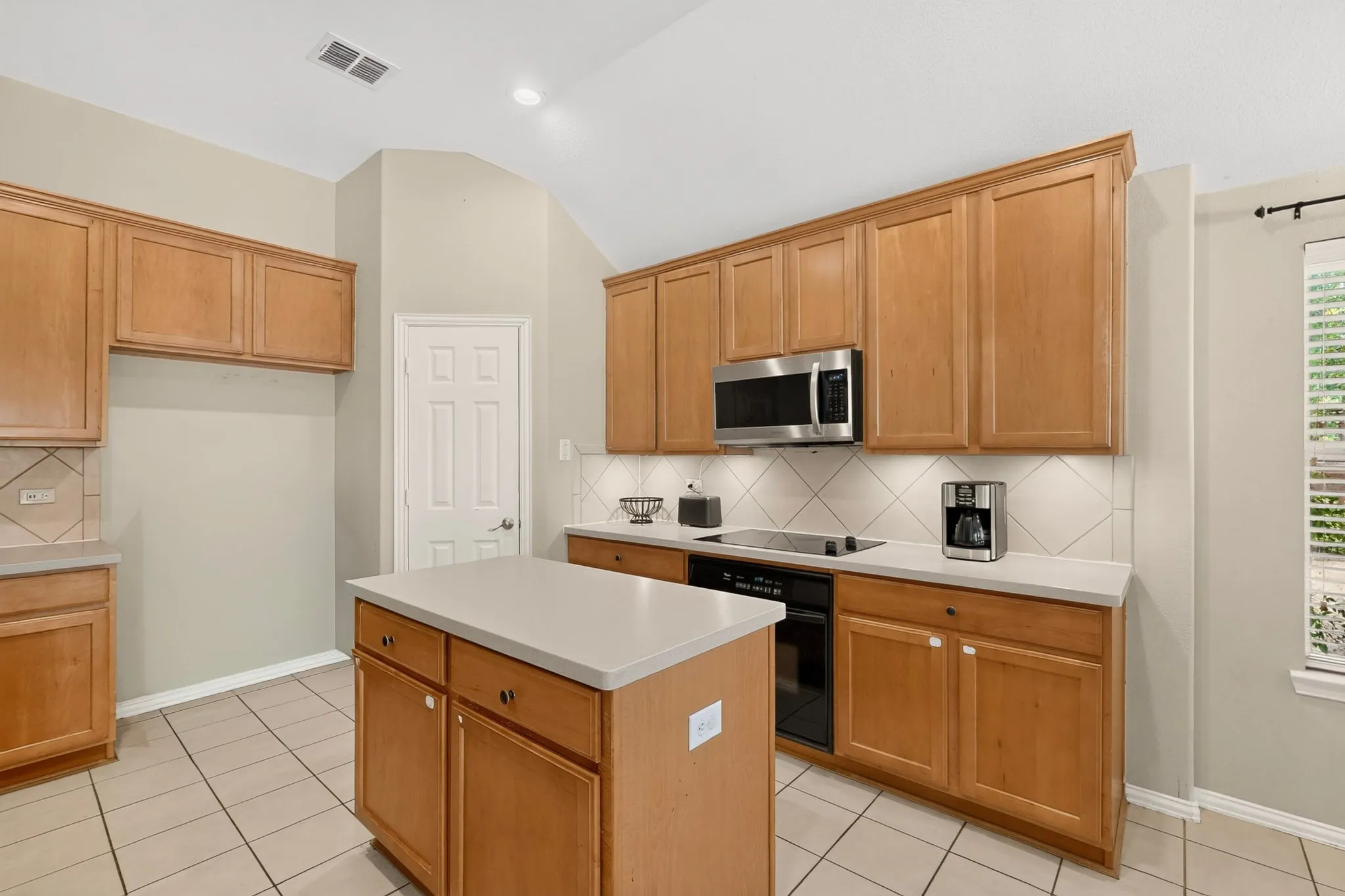 Kitchen featuring light wood cabinetry, a central island with a light countertop, stainless steel microwave, black cooktop, and tile flooring