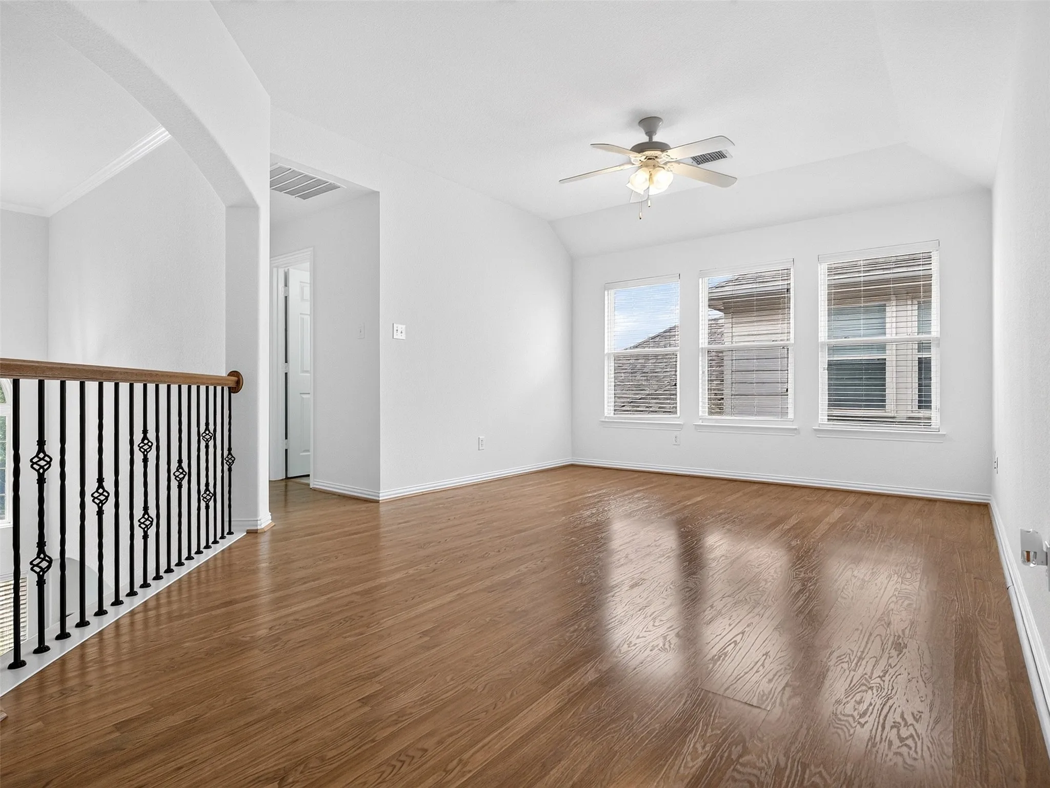 Wood-finish flooring, three windows with blinds, ceiling fan, and staircase with wrought iron balusters